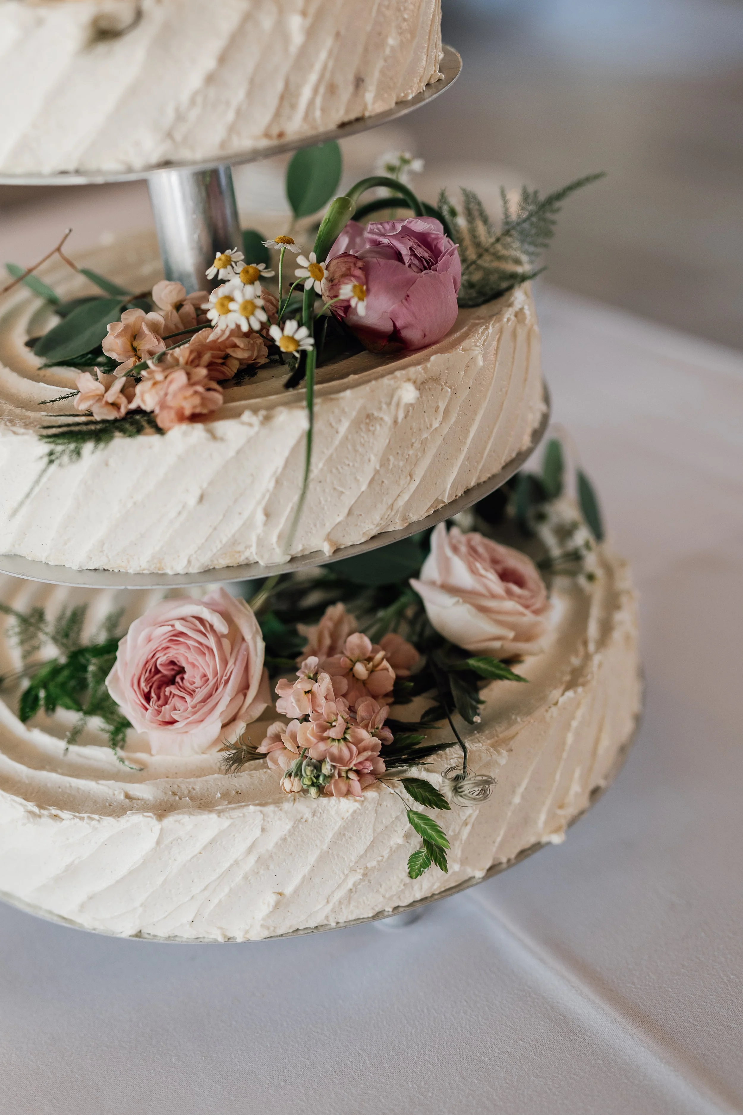 Nærbillede af en to-etagers bryllupskage dekoreret med pastelfarvede blomster, herunder rosa roser og små hvide blomster, med en neutral frosting og grønne blade.