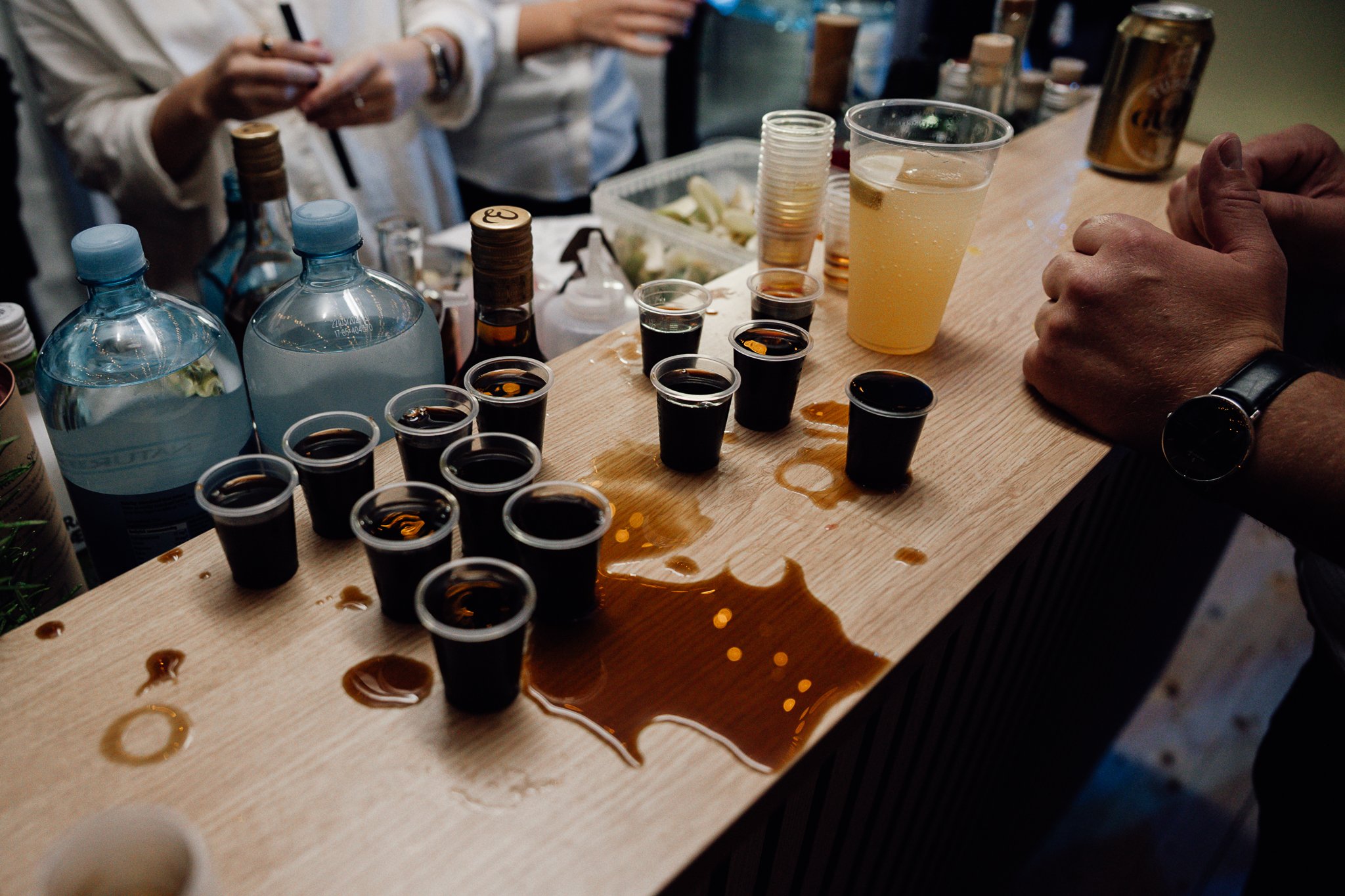 Nogle personer, der sidder ved en bar, med forskellige drikkevarer på bordet, herunder kobberkopper med mørk væske og en plastikkop med en gylden drik. Der er også nogle forskellige flasker og kopper i baggrunden.