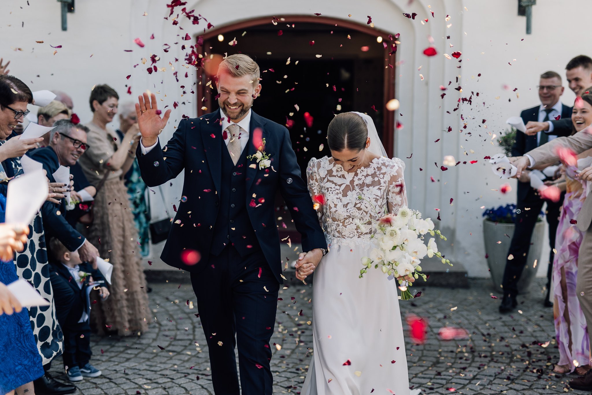 En brudepar går hånd i hånd ud af en kirke, mens gæster kaster blomster og konfetti rundt om dem. Manden har kort blond hår og griner, mens kvinden har mørkt hår og nedbøjet hoved, båret i en hvid blomstret brudekjole. Gæsterne smiler og har festlige