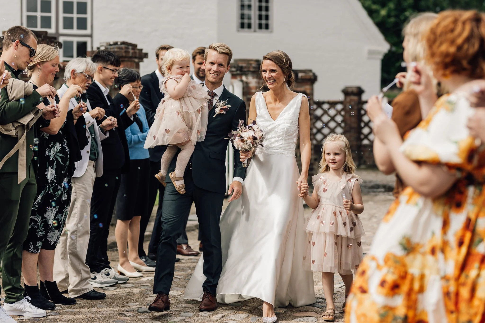 C: f8lling af et bryllup med brudepar og gæster, der sender bobler. Bruden b e6rer en hvid kjole, mens brudgommen har jakkes e6t og slipset. Sm e5 børn er til stede, og omgivelserne er udend e6rs med en hvid bygning i baggrunden.