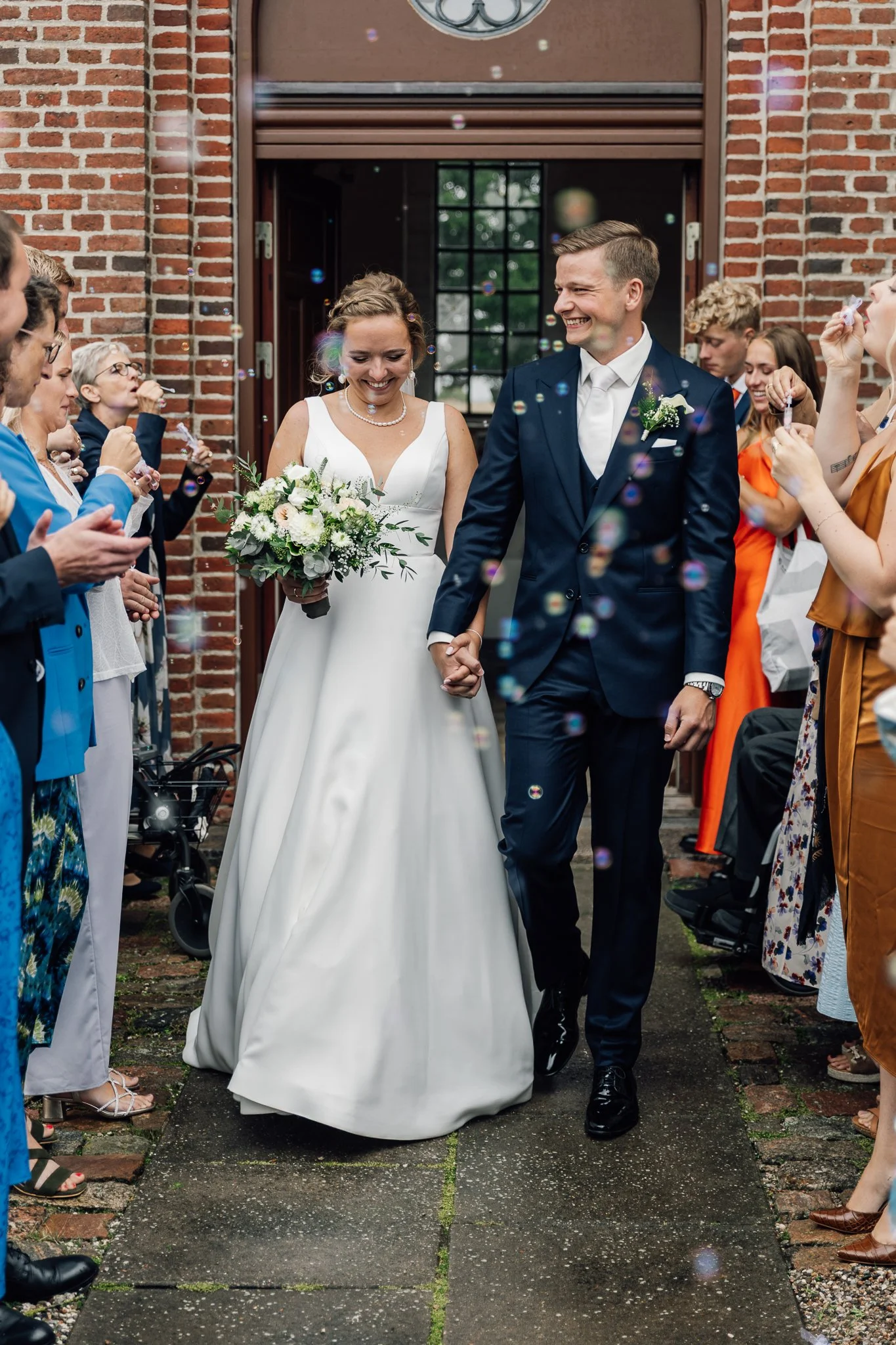 En brud og gommem foran en gruppe gæster udenfor en murstensbygning, holder hinanden i hånden og smiler, mens gæsterne kaster konfetti og røg.”}]]}″} 199}:{ 