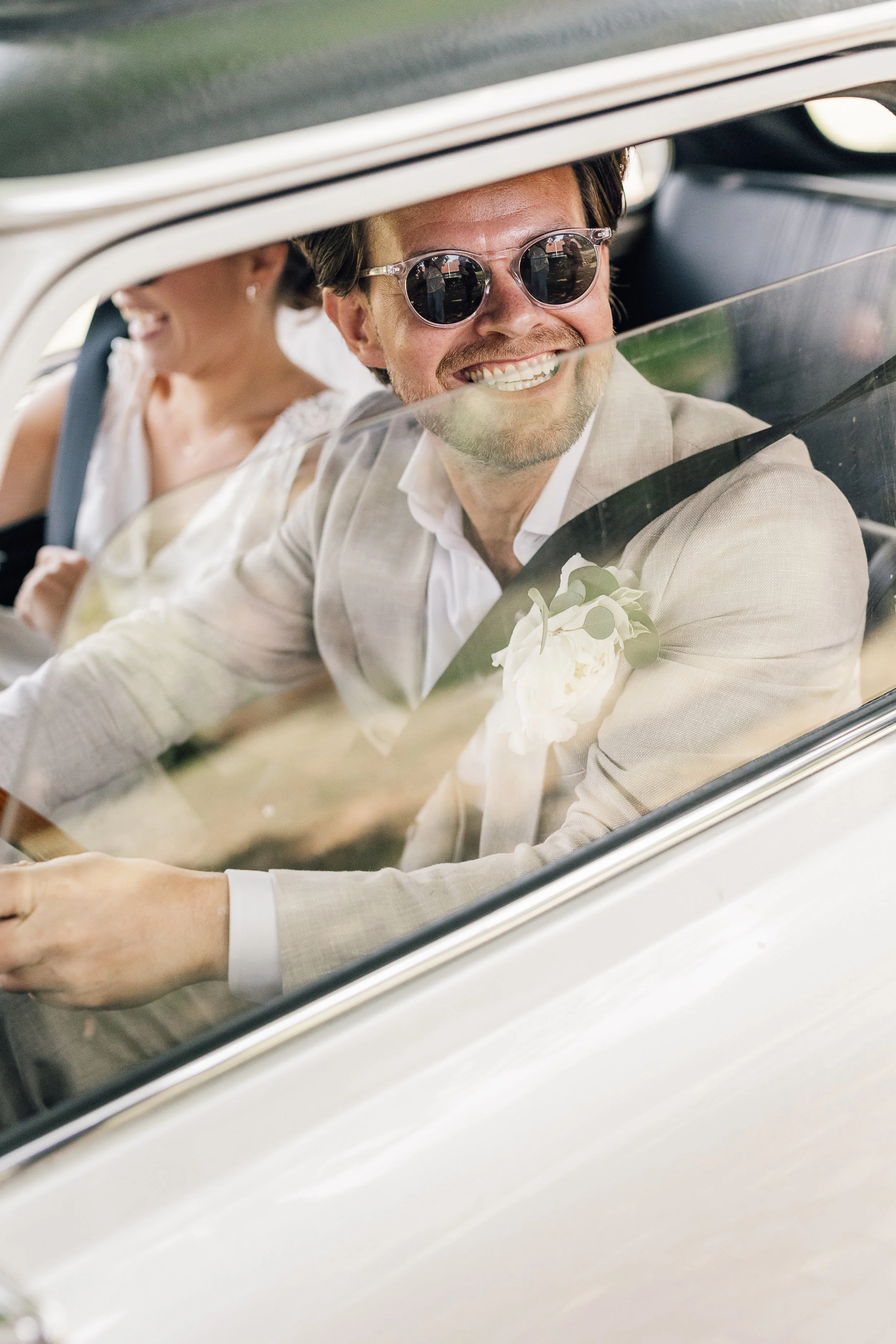 Glad mand og kvinde sidder i en bil, manden har sunglasses og en hvid jakke med en buket blomster på brystlommen, kvinden har en hvid kjole og sidder bagved.