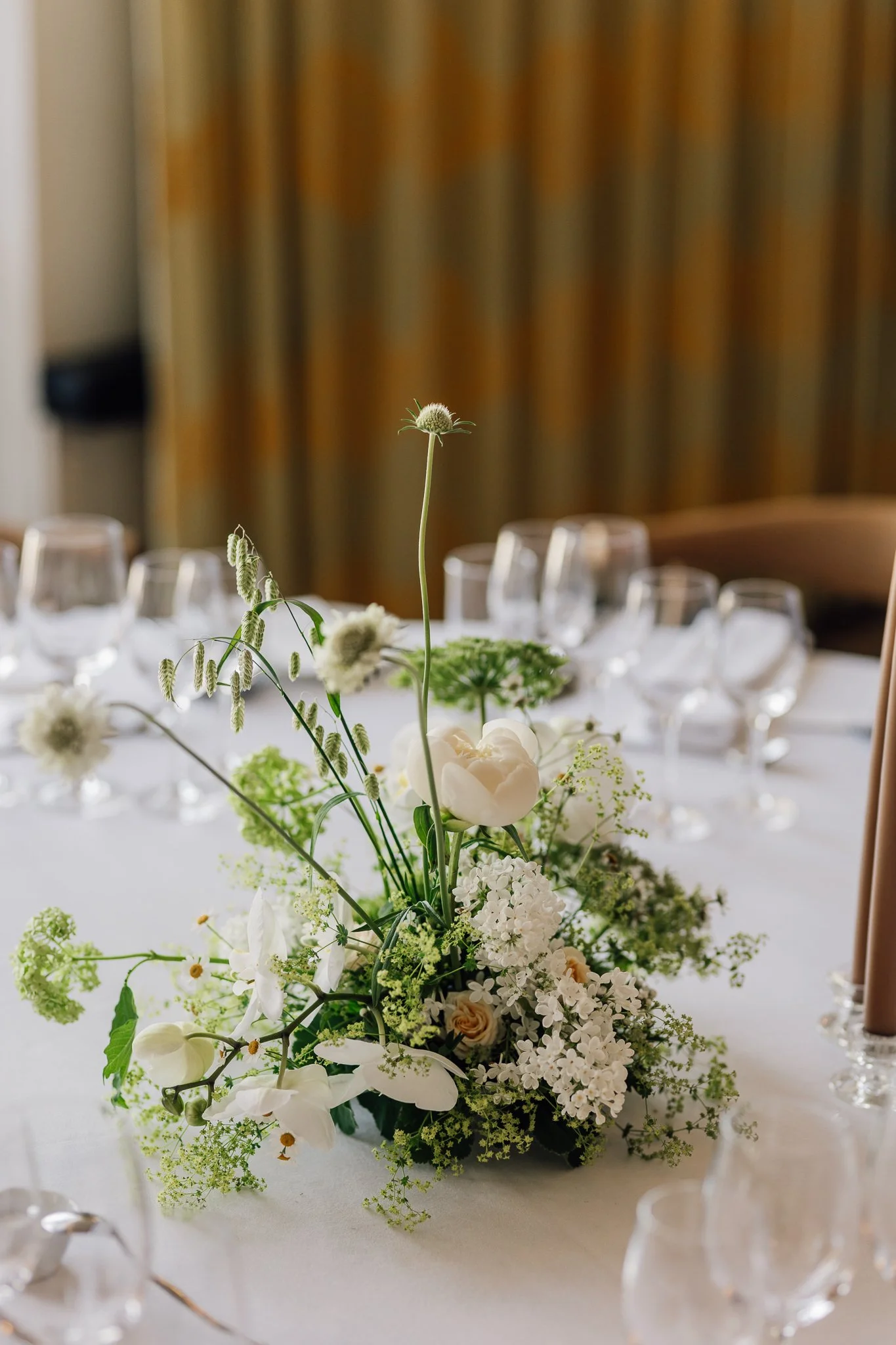 Buket af hvide og lyserøde blomster med grønne blade, placeret på et bord med glas og bestik, i et elegant indendørs festlokale.