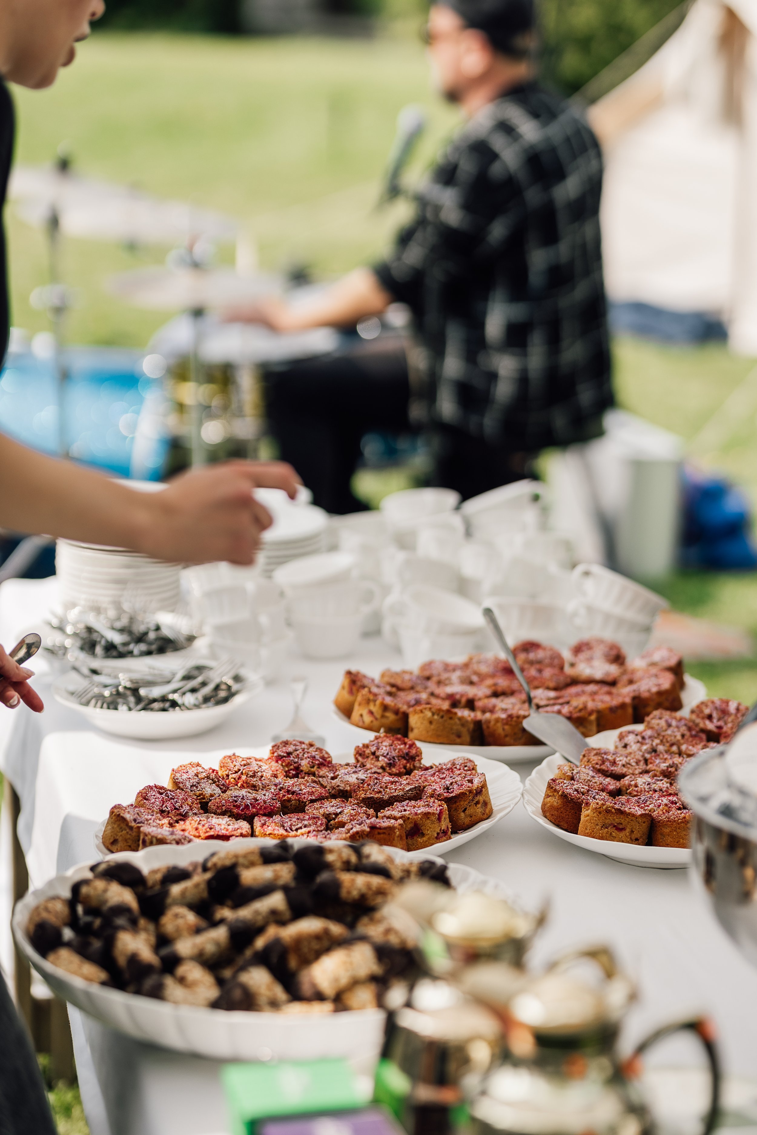 Udsigt over en buffet med kager, frugt og snacks ved en udendørs begivenhed med personer i baggrunden.