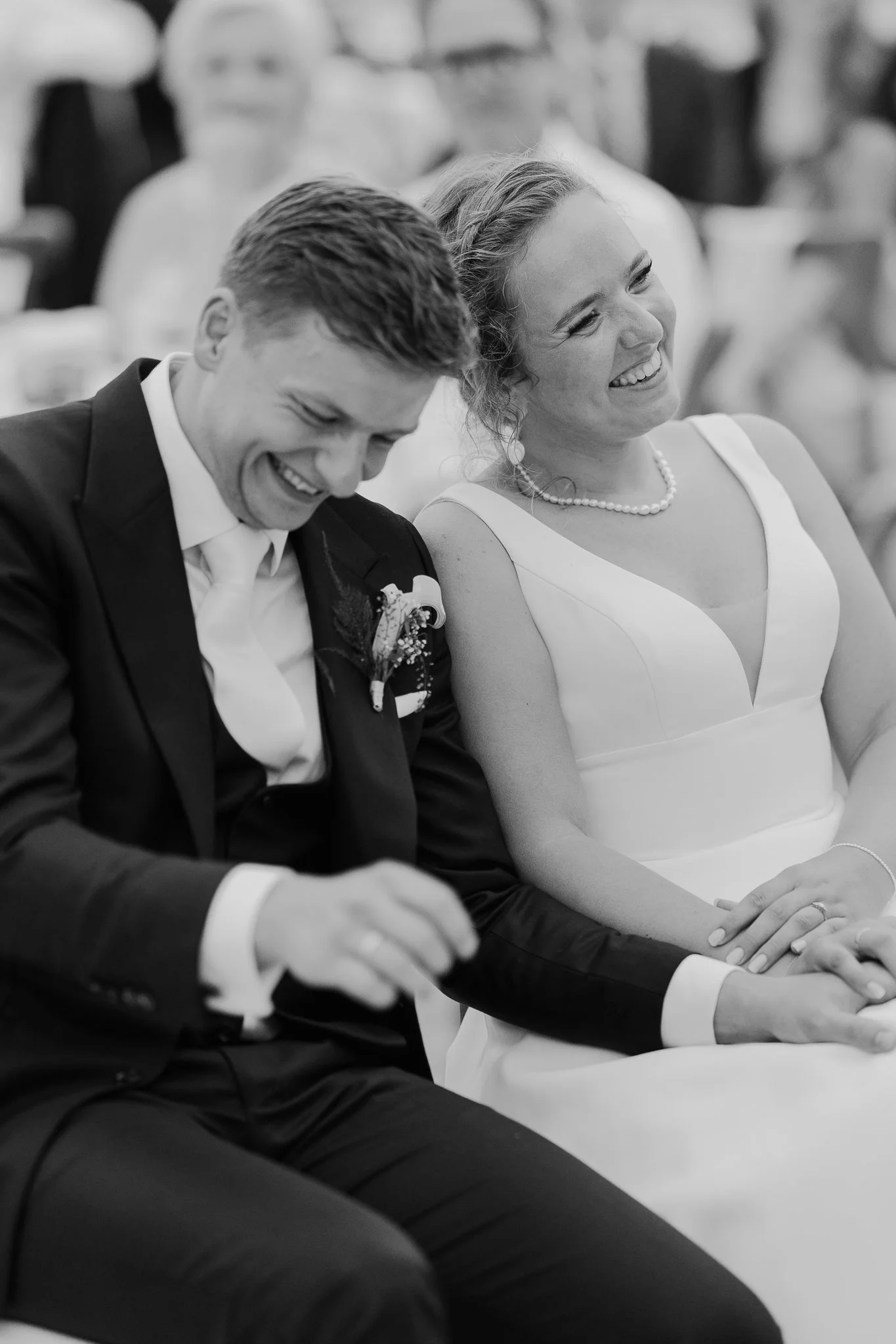 Et billede i sort-hvid af et lykkeligt brudepar, der sidder sammen ved en ceremoni, iført formelt tøj. Kvinden i hvid kjole, og manden i sort jakkesæt, begge er smilende.
