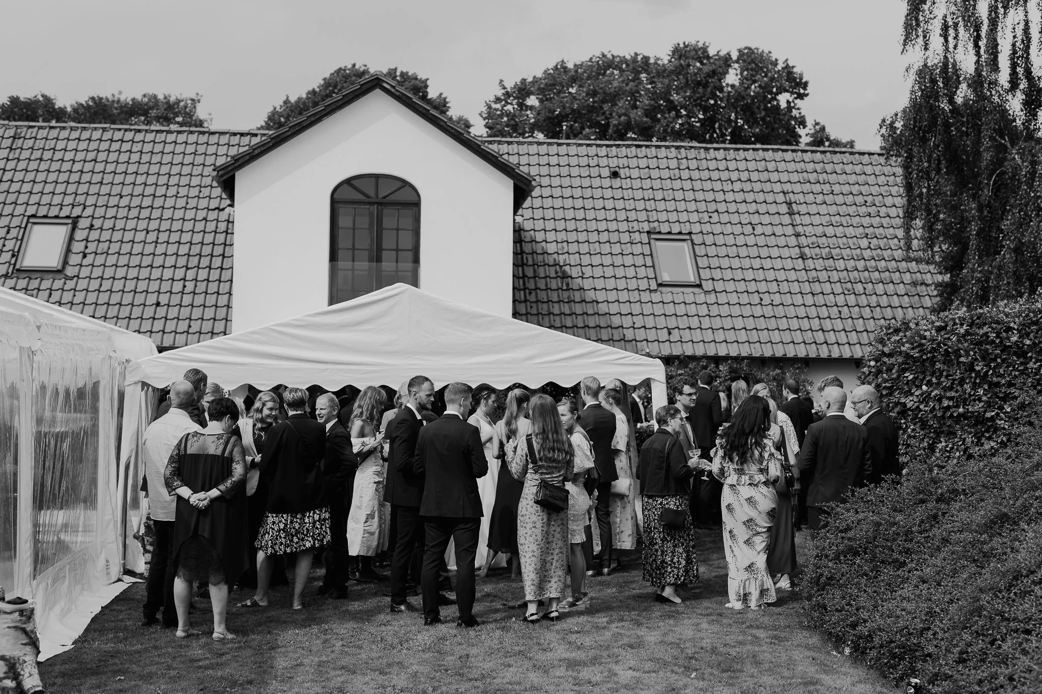 En gruppe mennesker samles udenfor foran en hvid bygning med skråt tag, til en begivenhed, under en hvid telt. Der er mange mennesker, der snakker og socialiserer.