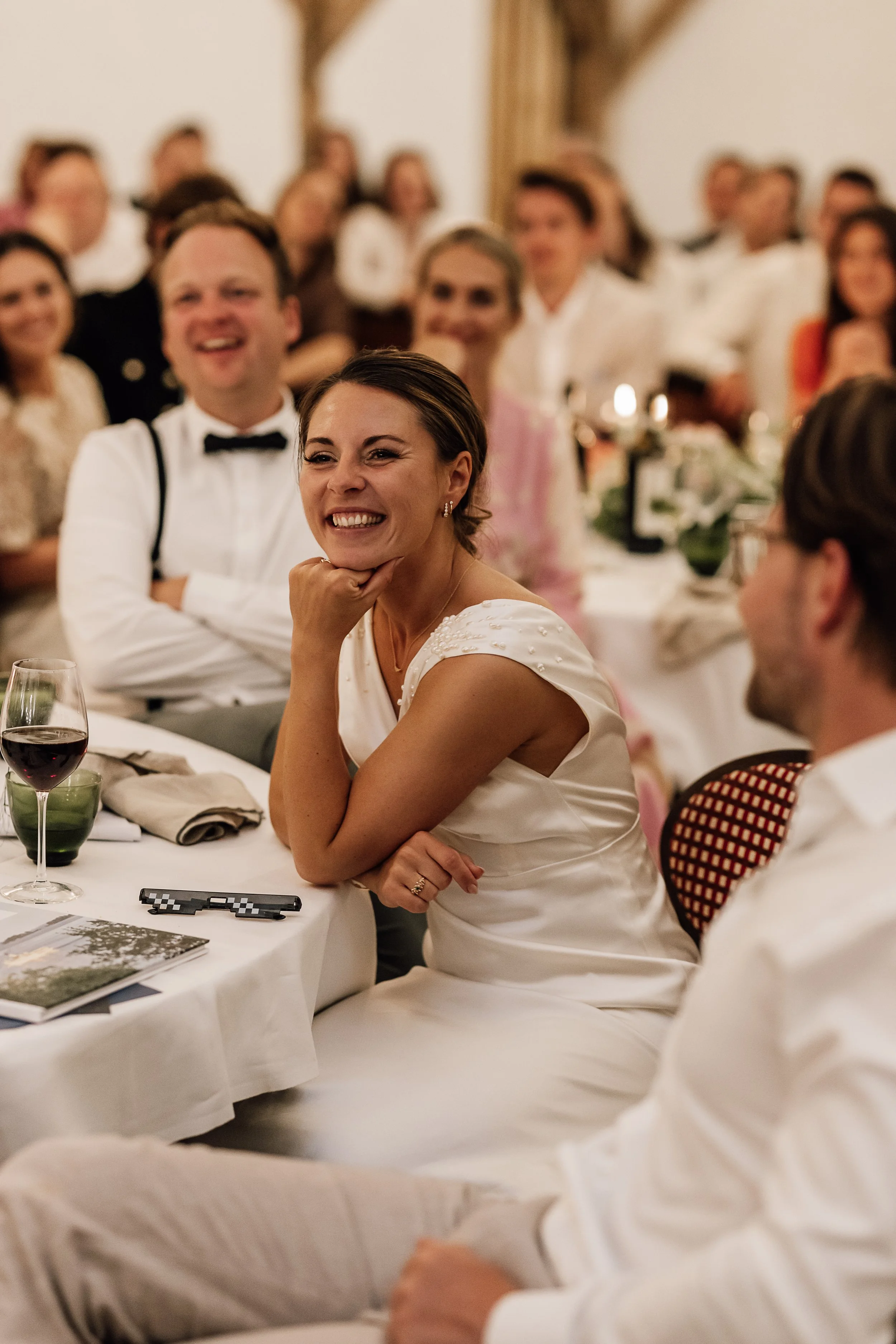 En gruppe mennesker til en formel middag, hvor en kvinde i forgrunden smiler og hviler sin hage på sin hånd, omgivet af andre personer, alle iført formelle kjoler og skjorter, i en elegant indretning.