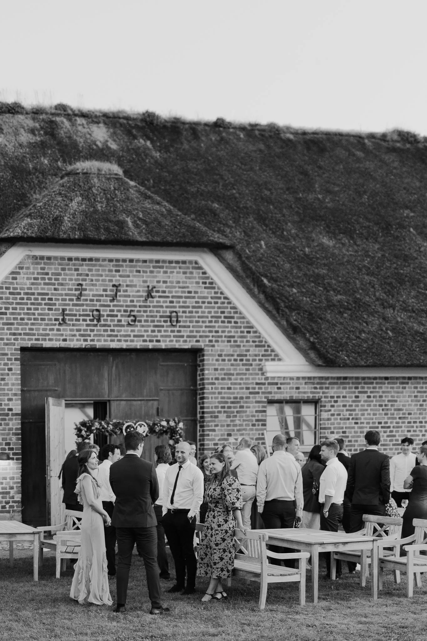 En gruppe mennesker, der ser ud til at være ved en fest udenfor en murstensbygning, hvor der er en dør dekoreret med blomster.