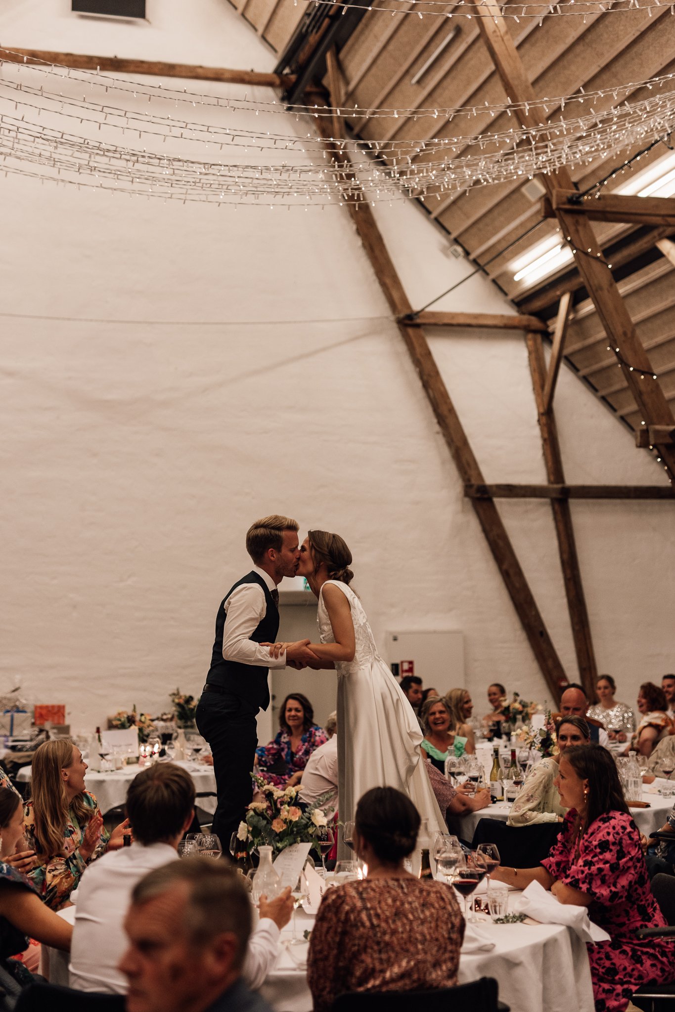 Et brudepar danser for et lykkeligt publikum ved et bryllup i en stor, træinddækket hall, med festlige borddekorationer og bløde belysning.