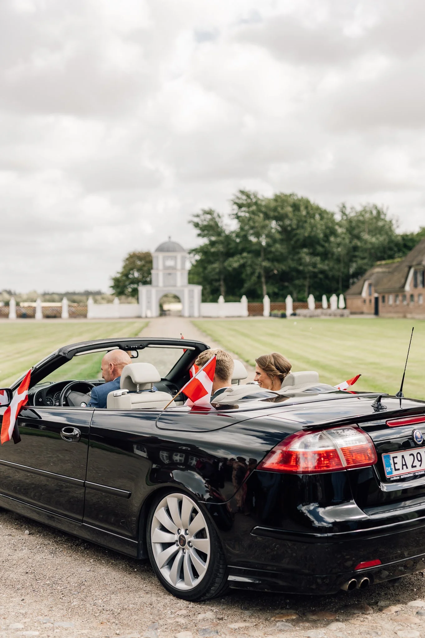 Sort bil med damer i cabriolet, dekoreret med små danske flag, kører på vej foran en historisk bygning med hvide spidser, omgivet af grønne træer og en græsplæne.
