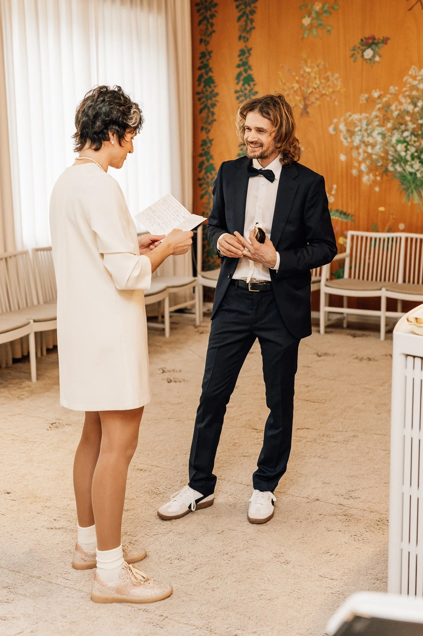 En mand i smoking og en kvinde i en lys kjole ved et bryllup, hvor kvinden læser en tale. De er inde i en indendørsbryllupslokale med blomsterdekorationer på væggen.