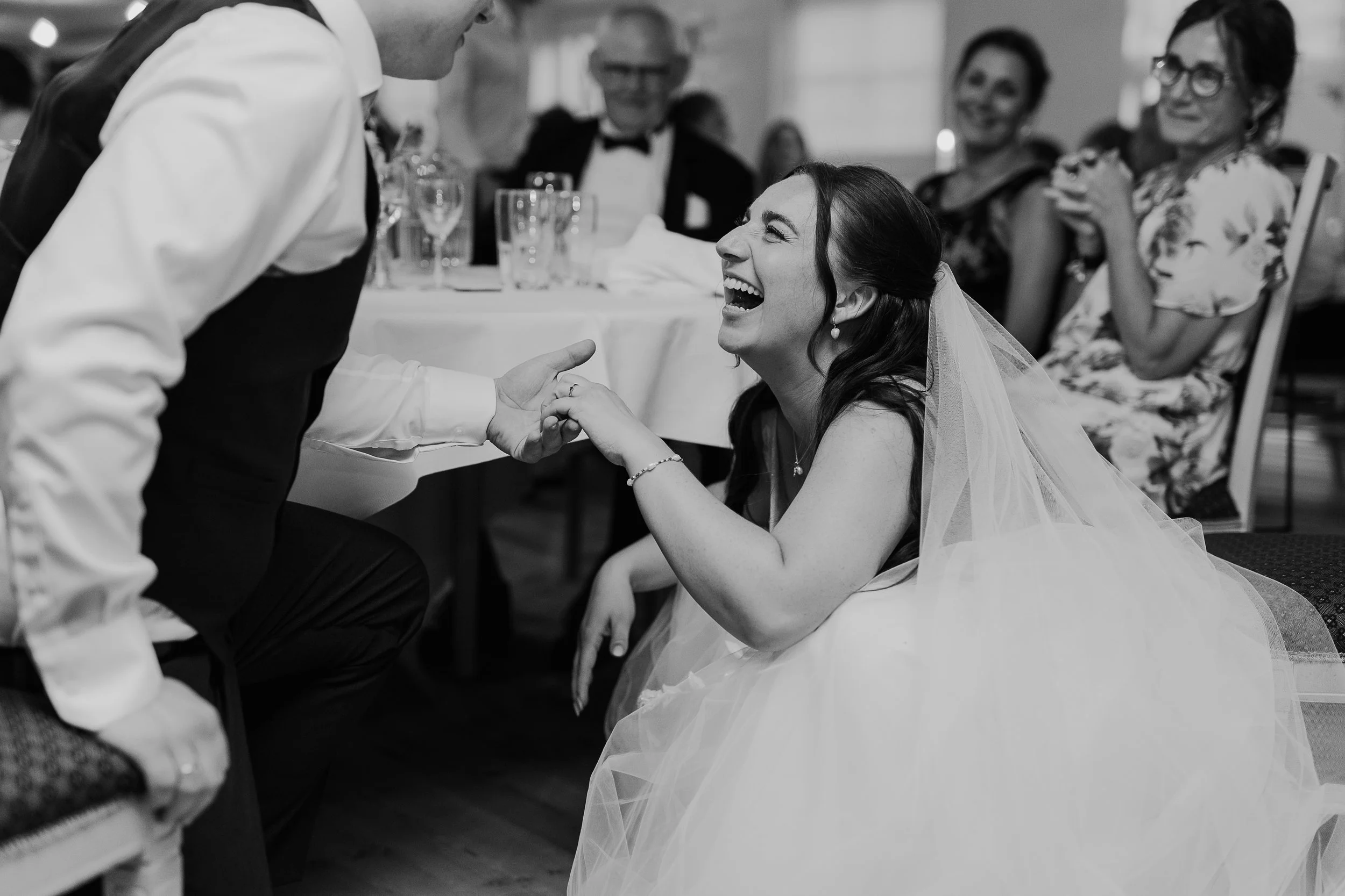 Sort-hvid foto af en bryllupsscene, hvor en brud og en gæst holder i hånd og smiler. Gæster i baggrunden sidder ved et bord og ser glade ud.