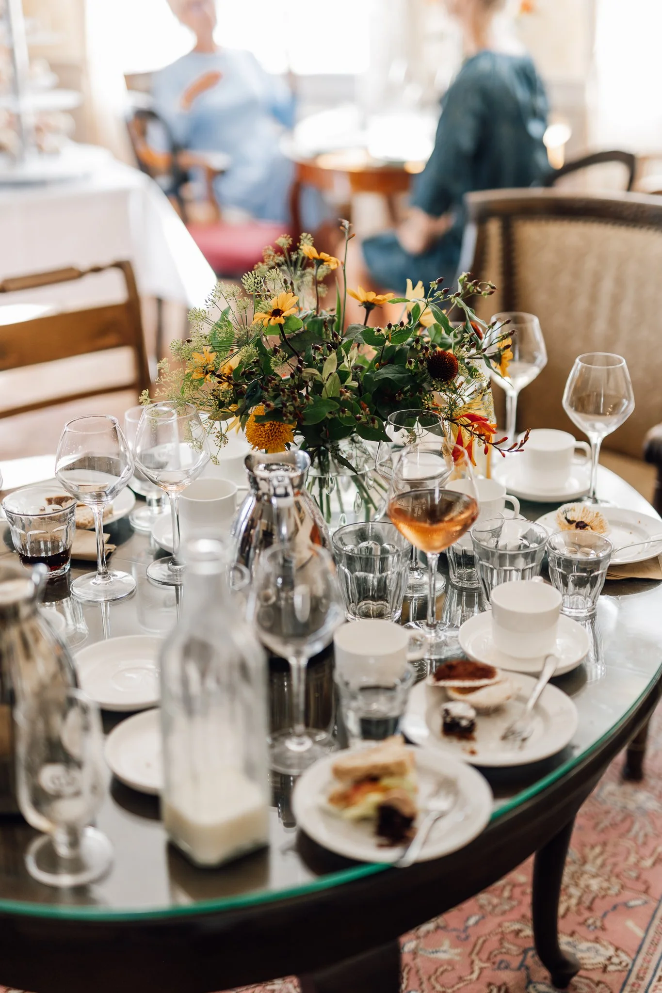 Bord med en blomsterdekoration, glas, tallerkener og rester af mad, på et festbord i en hyggelig spisestue. Der er to personer i baggrunden, der sidder og taler.