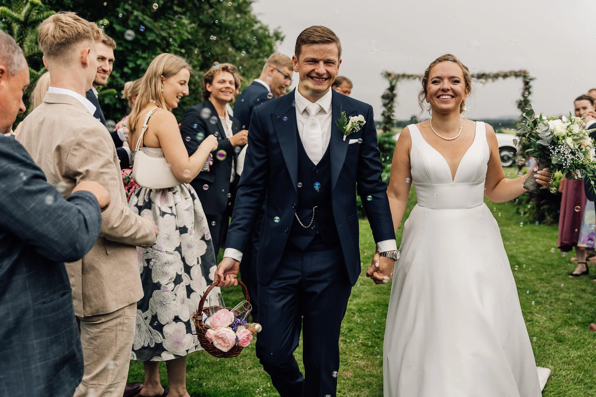 Et par bliver gift, omgivet af venner og familie, i en udendørs bryllupsceremoni med grønne træer og en blomsterbue i baggrunden.