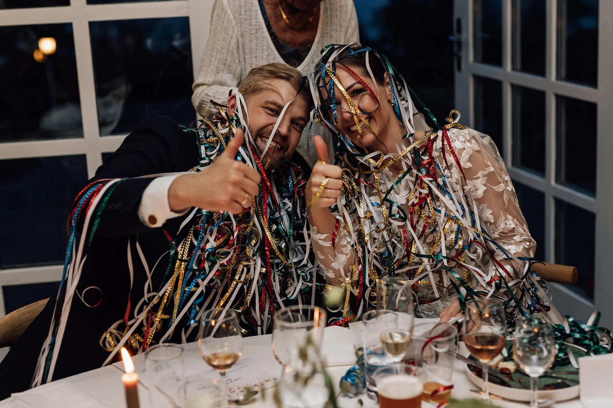 To mennesker sidder ved et festbord, overvundet med farverige sløjfer og konfetti, og smile og giver tommelfingre op. Der er vin i glassene på bordet. Bagved står en person, der også er glad.