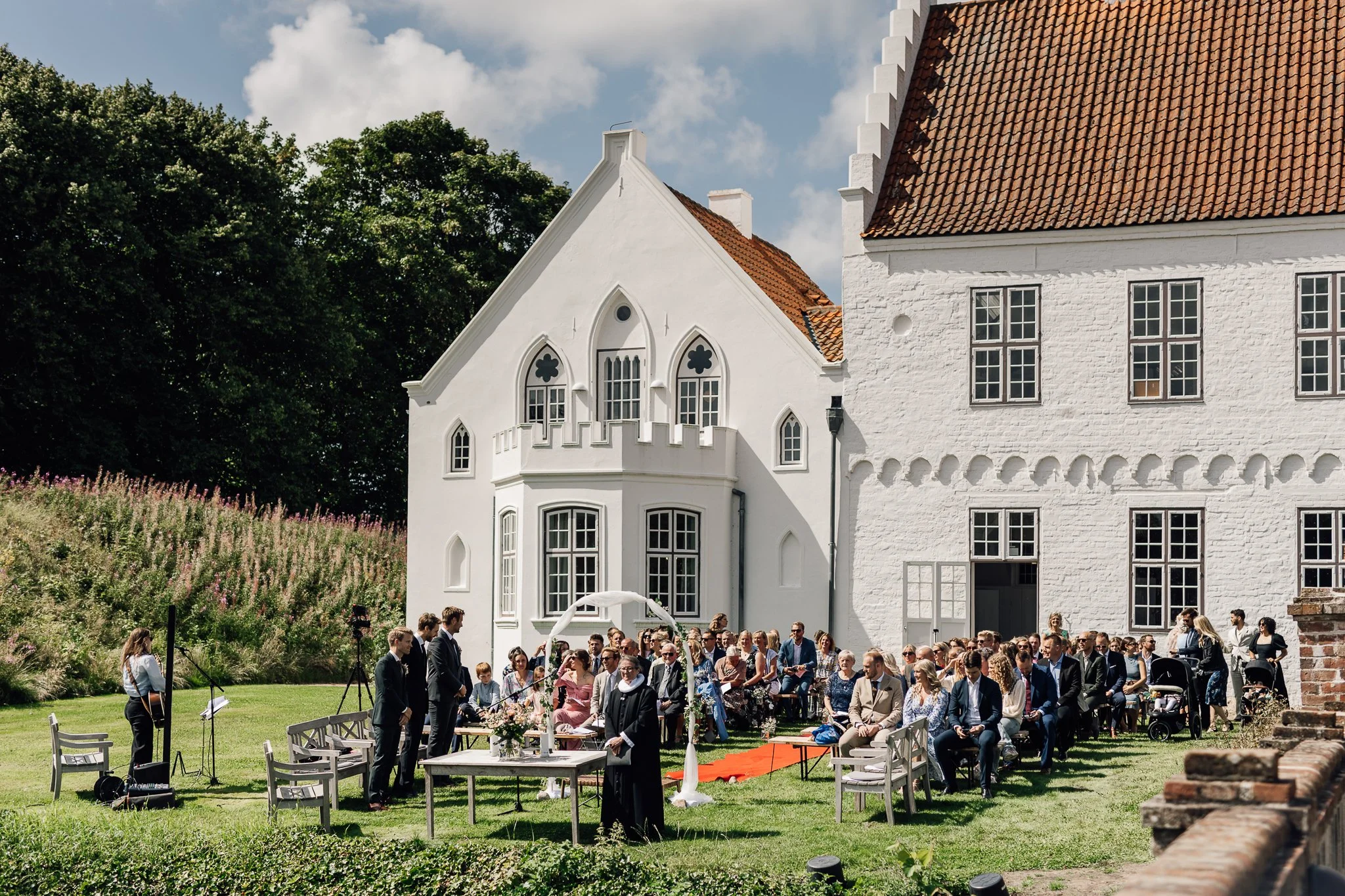 En stor gruppe mennesker er samlet udenfor foran en hvid, gammeldags bygning med rødt tegltag, sandsynligvis til en seremoni eller fest.