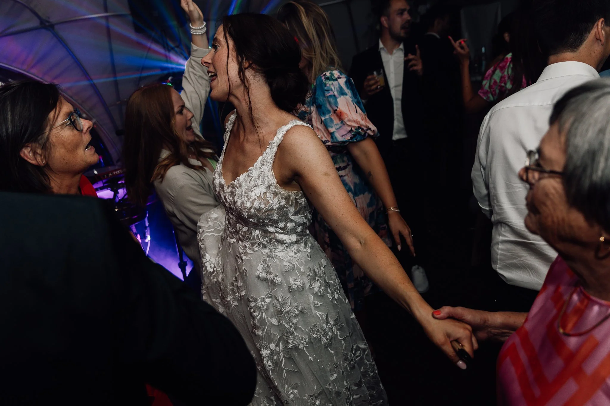 En gruppe mennesker danser og har det sjovt på en fest, med betoning på en kvinde i en hvid kjole, der smiler og holder en anden persons hånd.