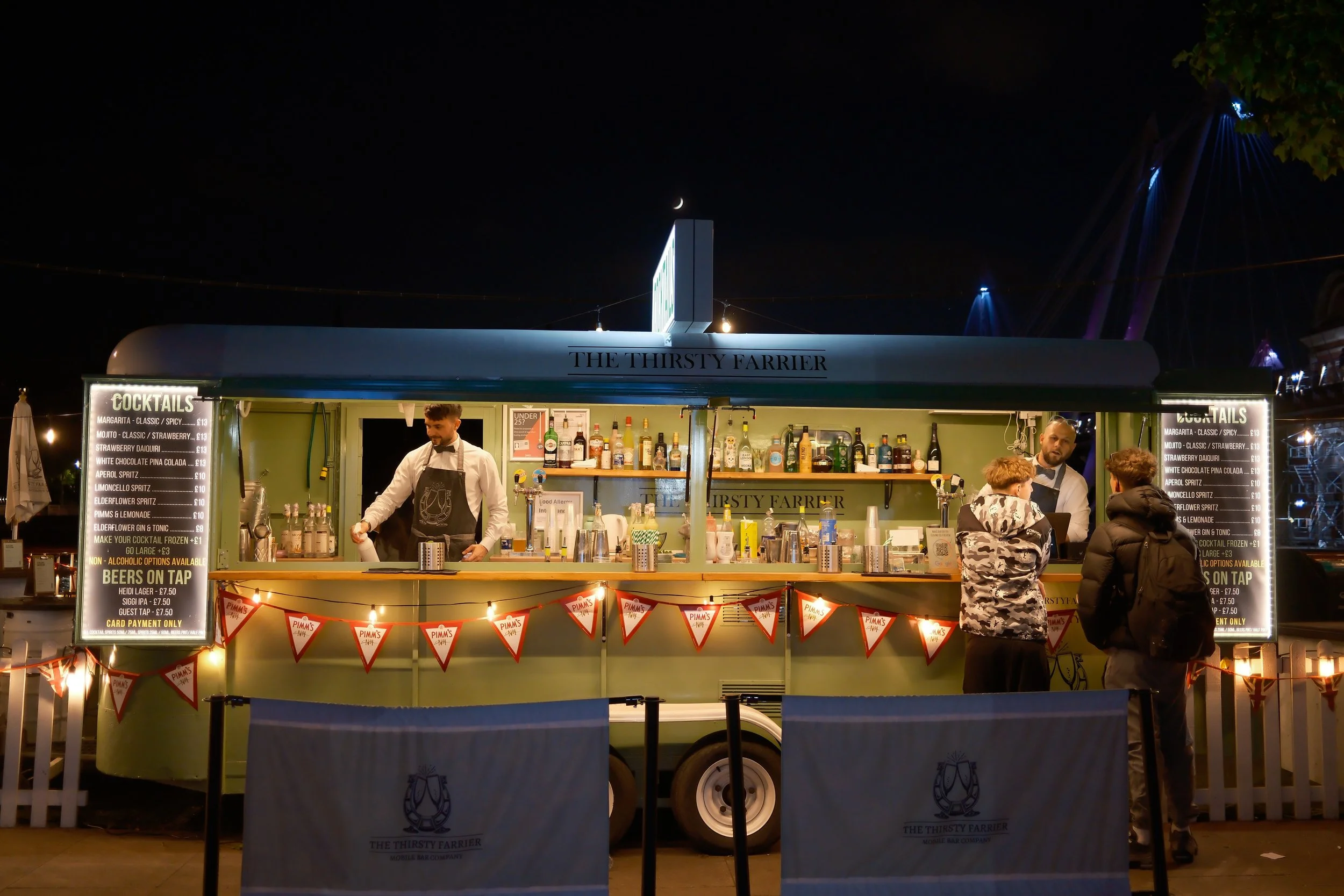 Night scene of a mobile bar named 'The Thirsty Farrier' with bartenders serving drinks at an outdoor event. The bar has a menu listing cocktails and beers, with colorful string lights, and a dark sky featuring a crescent moon and a faint star.