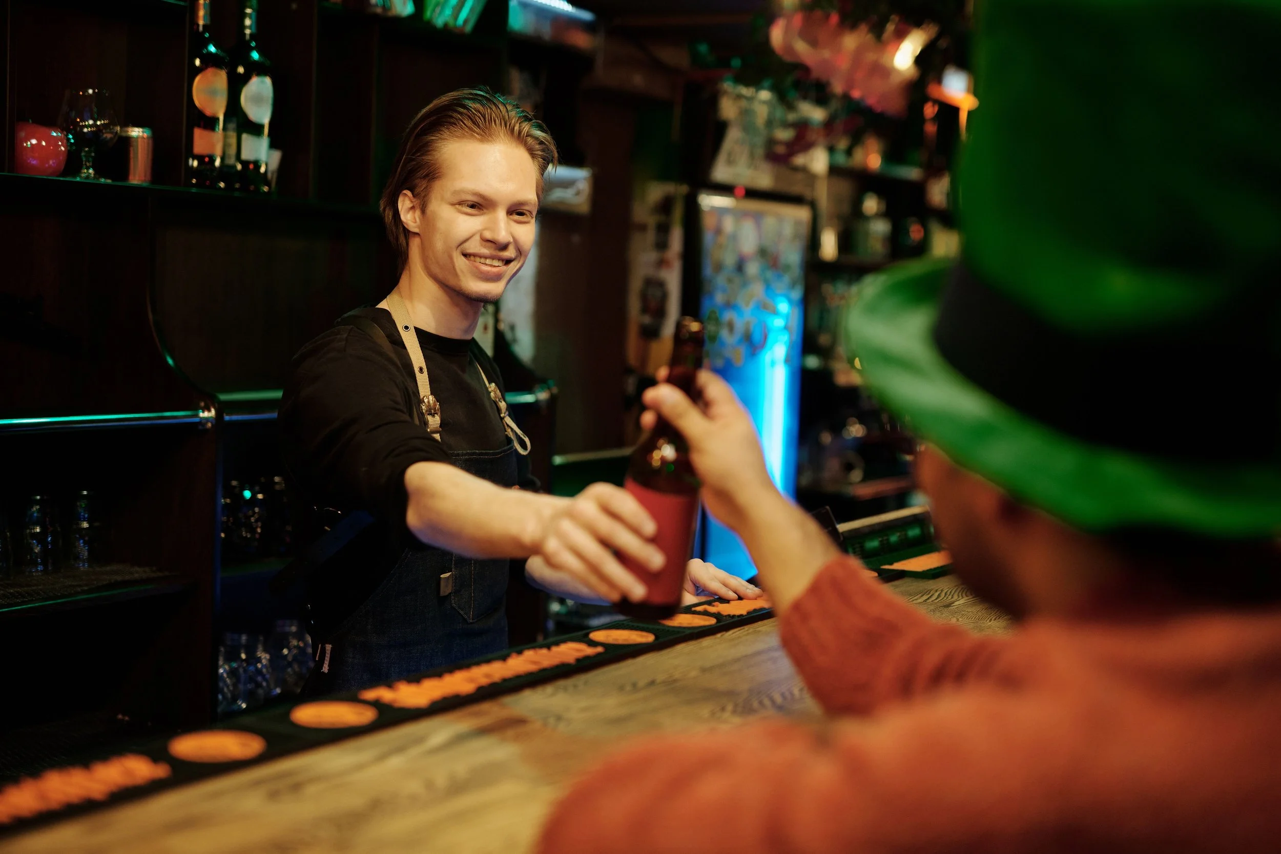 A bartender with short hair wearing a black shirt and apron smiling as they serve drinks to a customer wearing a green hat in a dimly lit bar.