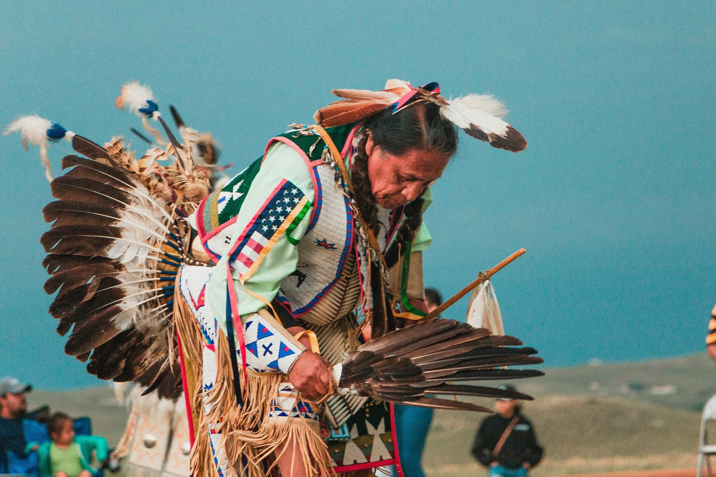 A Native American man dressed in traditional attire with feathered headdresses and beaded clothing, performing a cultural dance outdoors, with a clear sky in the background.