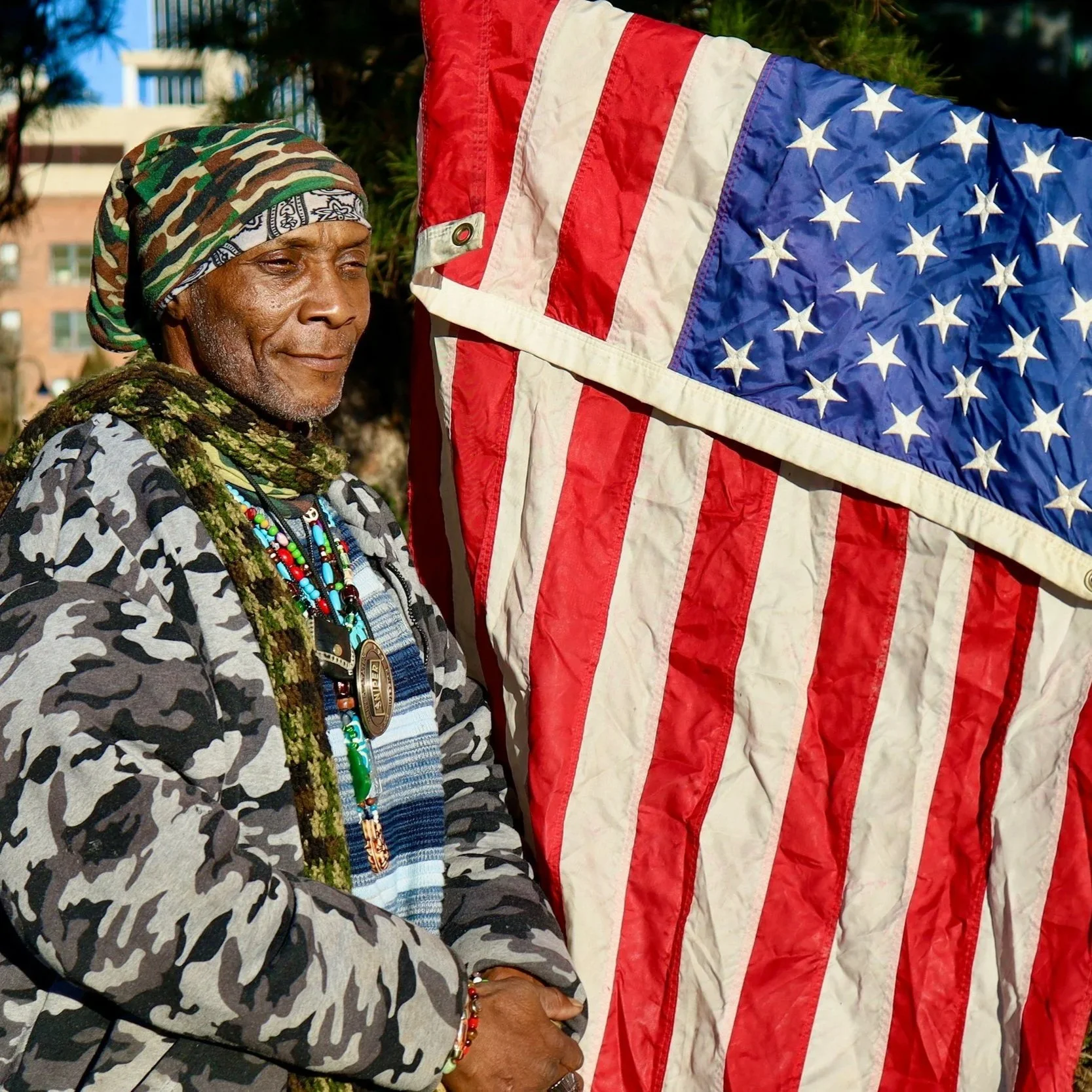 An elderly person wearing a camouflage headscarf, layered necklaces, and a camouflage jacket stands next to a large American flag displayed outdoors, with a background of trees and buildings.