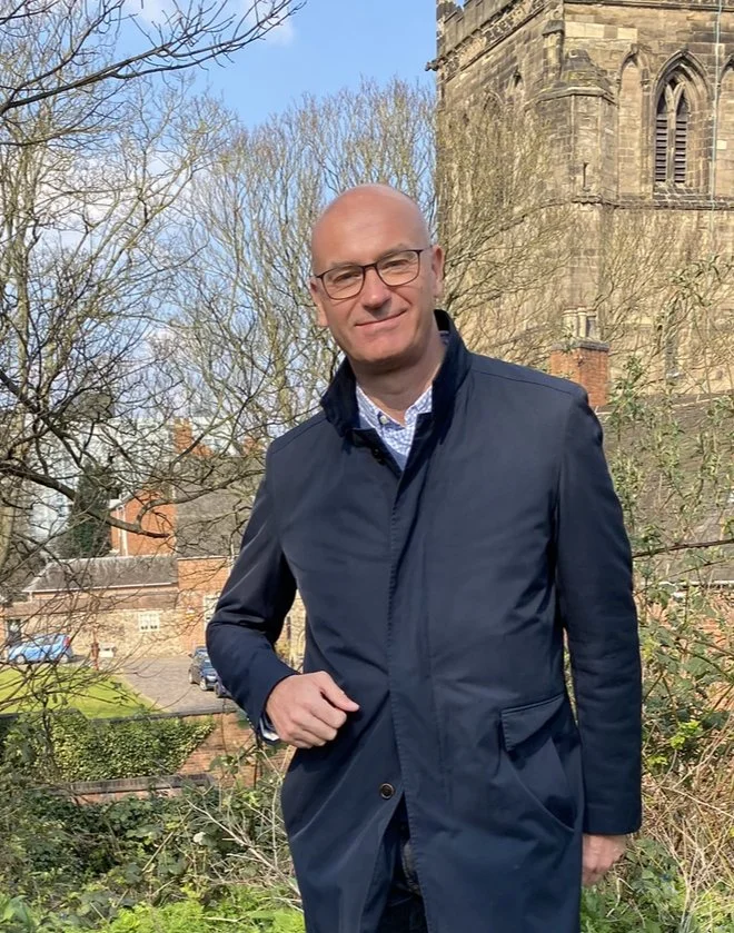 A bald man with glasses wearing a navy-blue coat, standing outdoors near a historic stone building, with leafless trees and parked cars in the background.