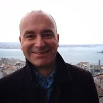 A smiling man with a bald head, wearing a dark jacket and blue shirt, standing outdoors with a cityscape and water in the background.