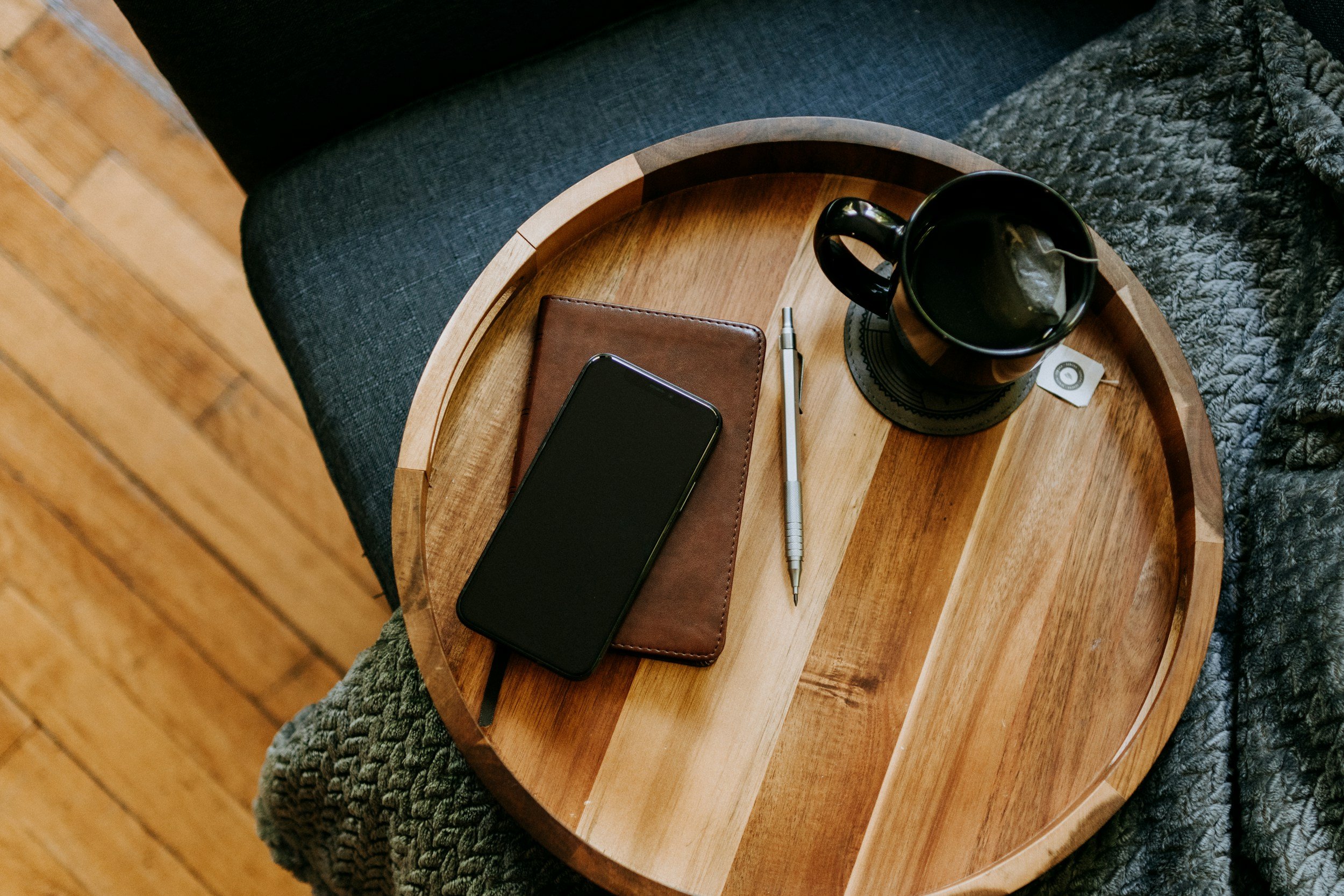 A wooden round coffee table with a black mug, a smartphone, a brown notebook, a silver pen, and a tea bag tag on it, placed on a gray knitted blanket near a gray upholstered armchair.