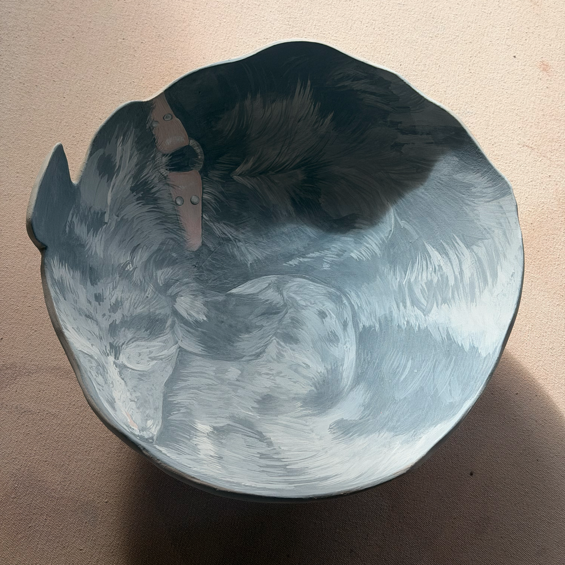 Close-up of a ceramic bowl with a painted image of a sleeping dog's face, lying on a brown fabric surface.