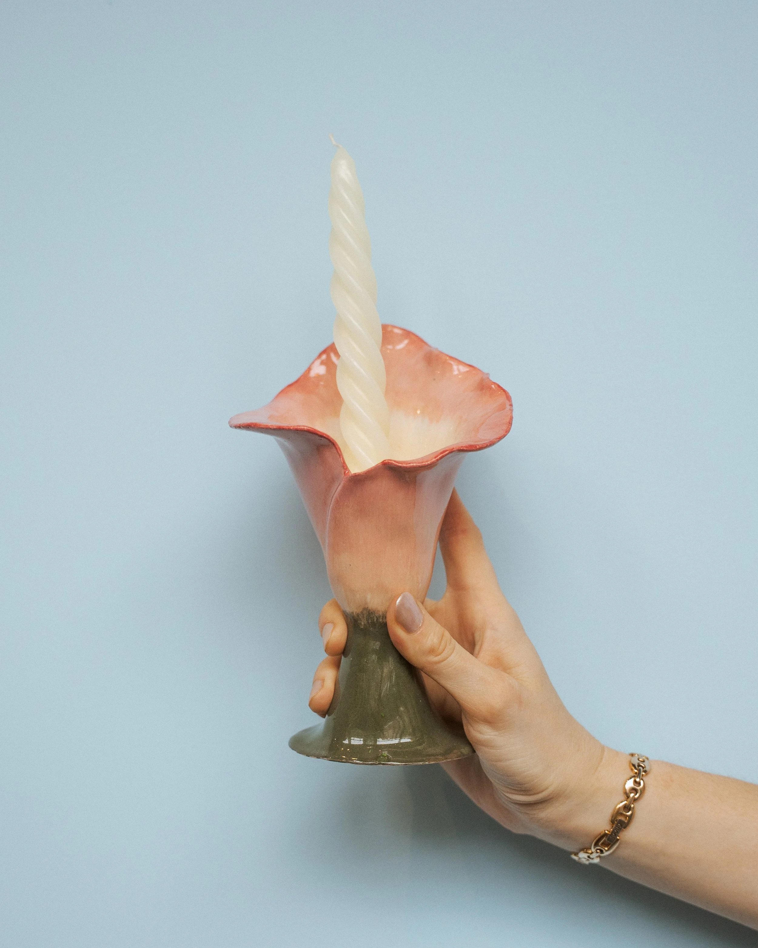 A hand holding a ceramic candle holder with a single pillar candle and a decorative flower-shaped holder. The hand is wearing a gold bracelet.