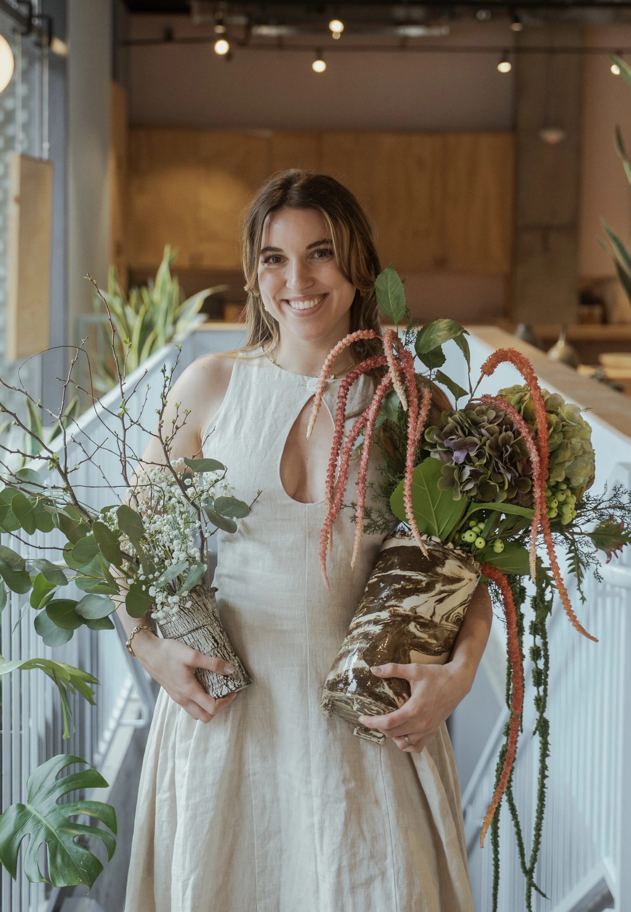 A woman with shoulder-length brown hair smiling, holding two large floral arrangements in a modern indoor space with plants and warm lighting.