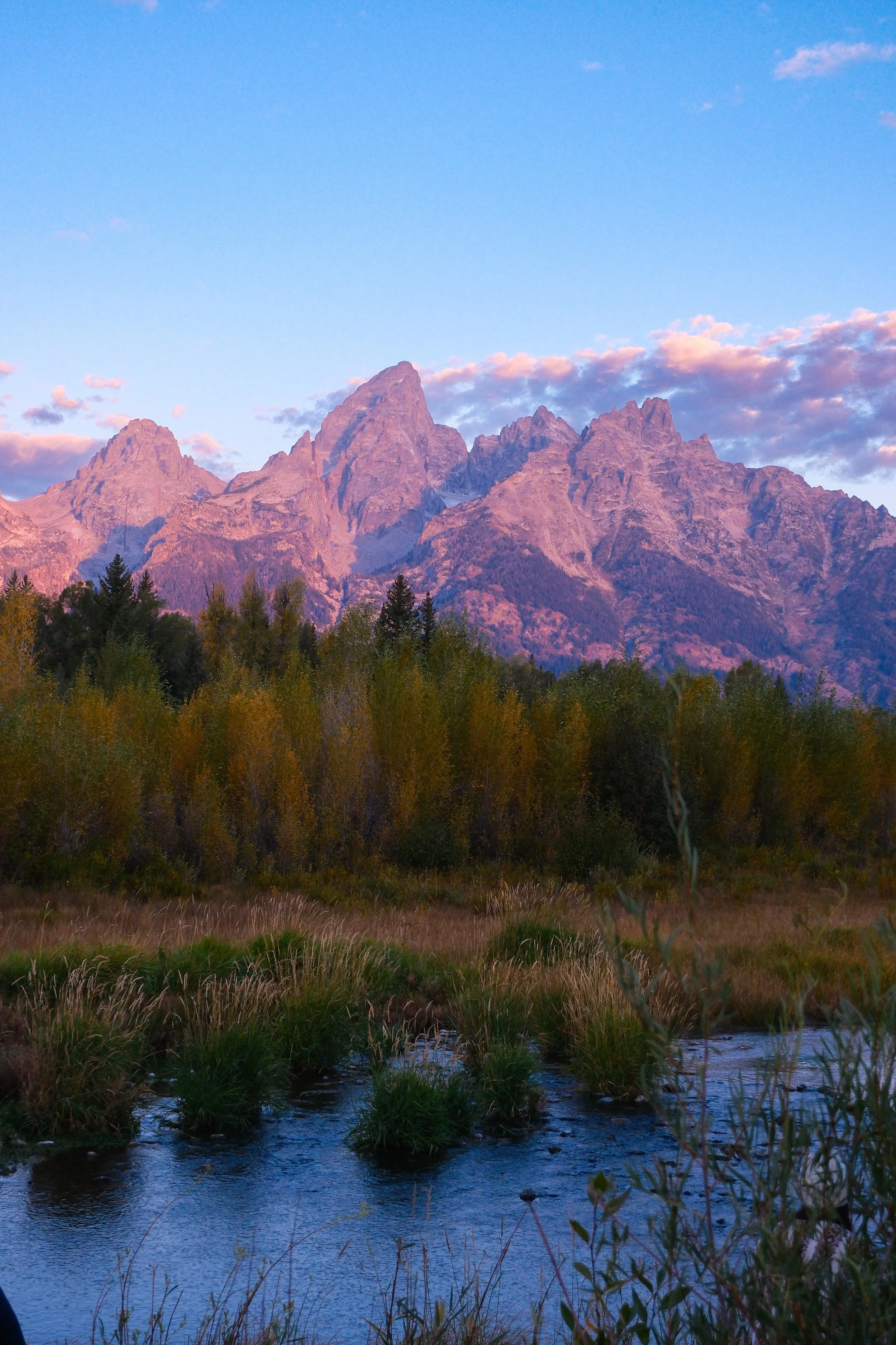 Mountain range at sunset with a river in the foreground and trees along the riverbank.