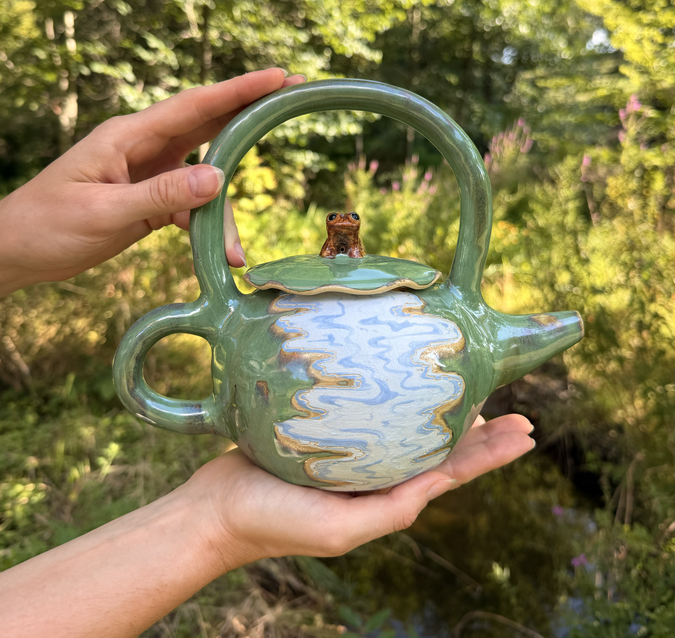 A ceramic teapot with a small frog figurine on the lid, held by two hands outdoors with a background of greenery and pink flowers.