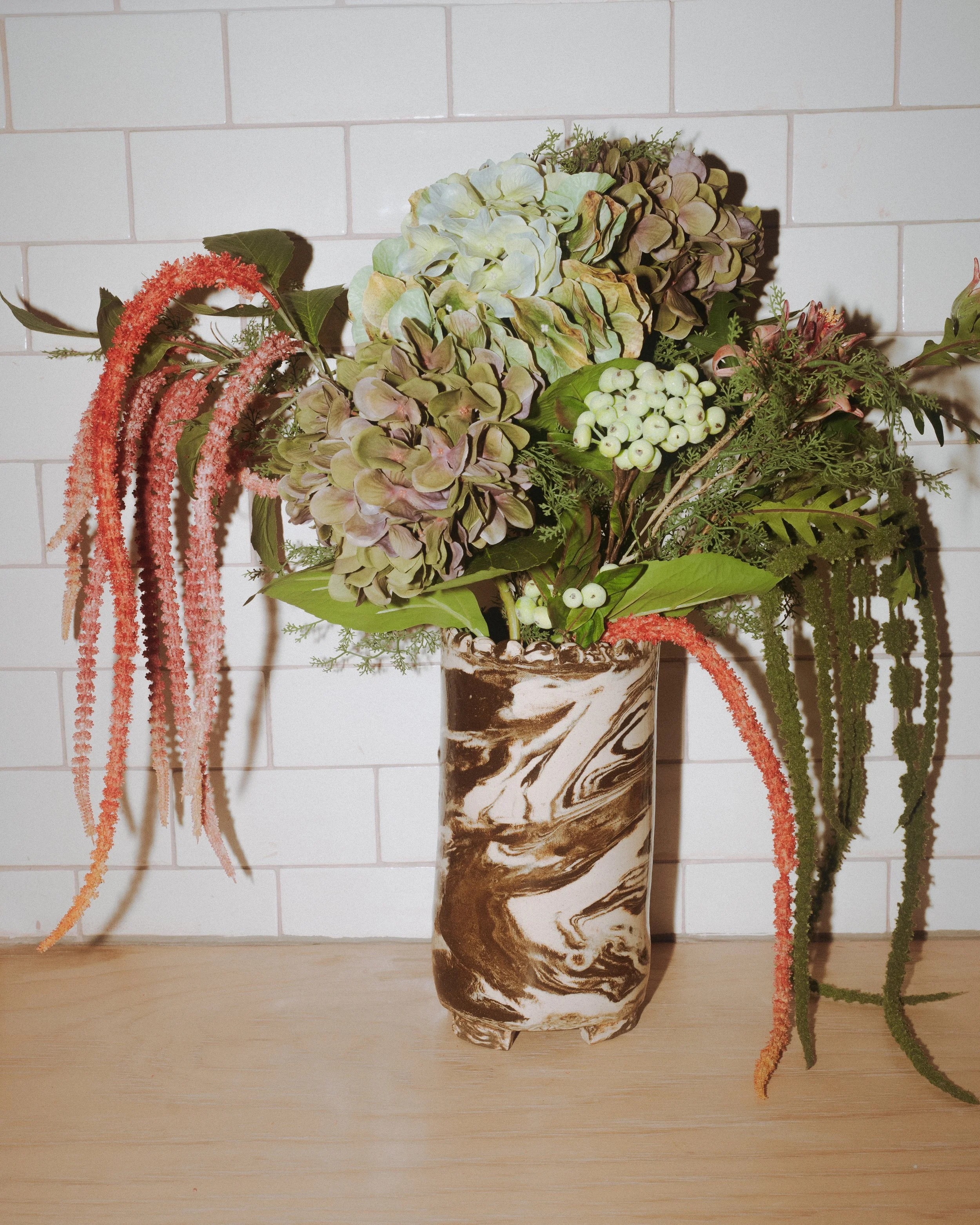 A tall marbled vase with swirling brown and white patterns on a wooden surface, filled with a mixed arrangement of dried and artificial flowers and greenery, set against a white tiled wall background.
