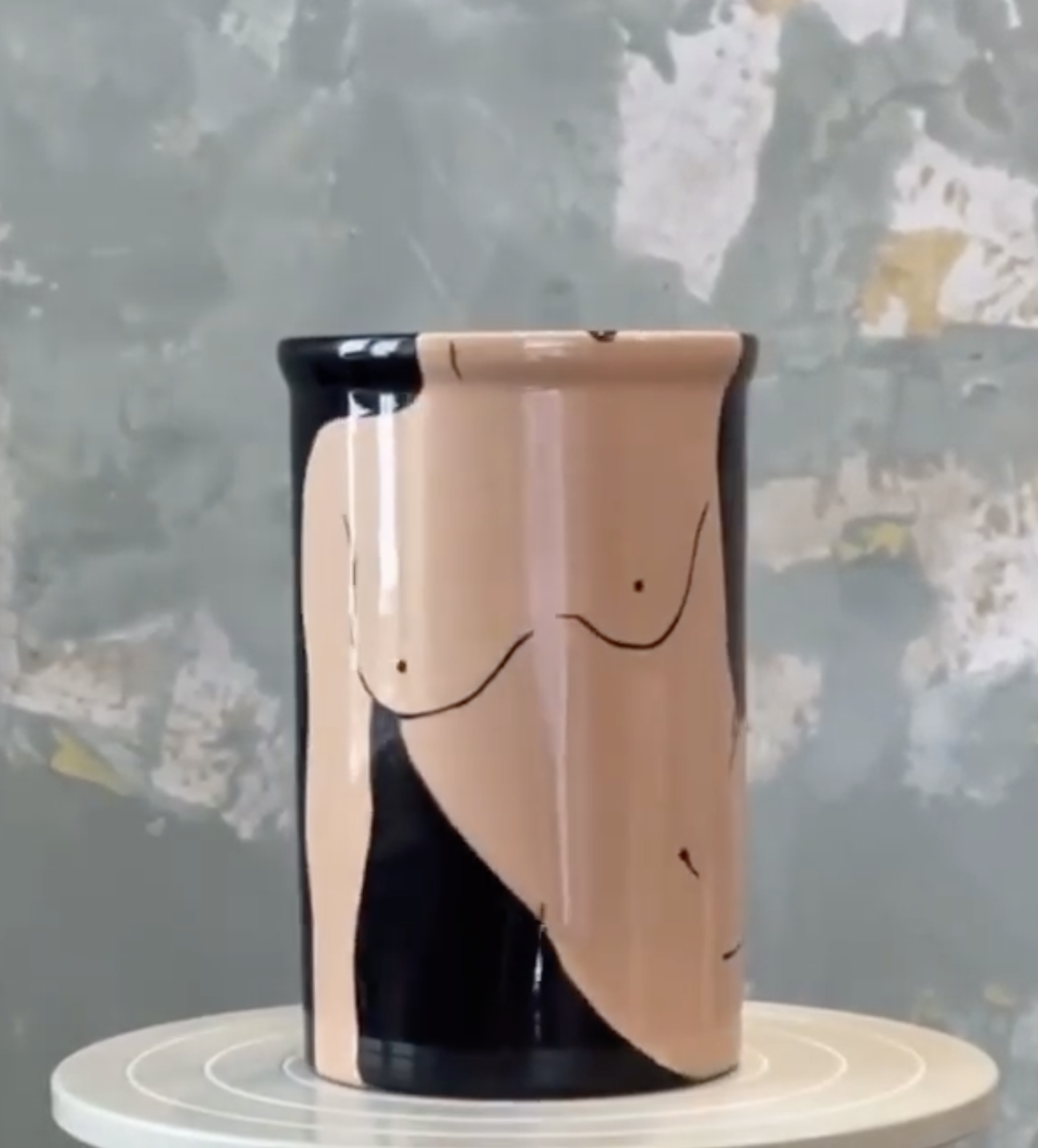 A biodegradable coffee cup with a minimalist black line drawing of a woman's chest on it, placed on a white circular platform.
