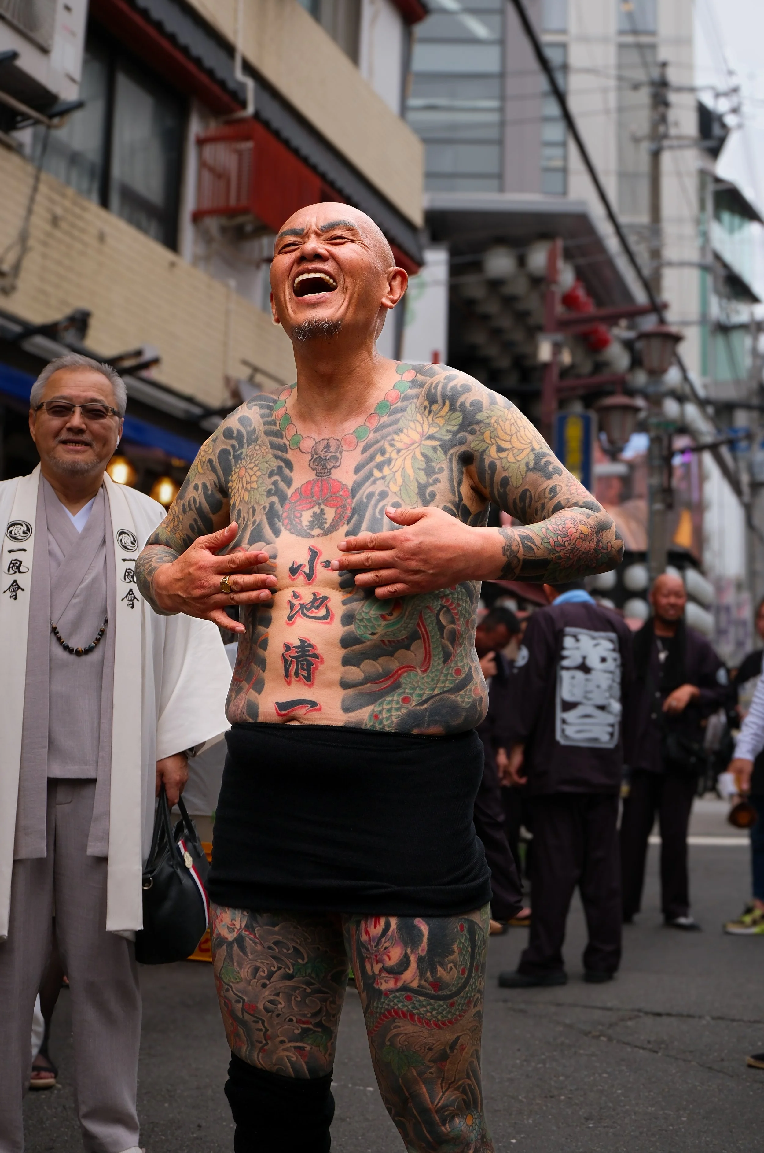 A bald, heavily tattooed man laughing with his eyes closed, standing shirtless in a street with people and buildings around.