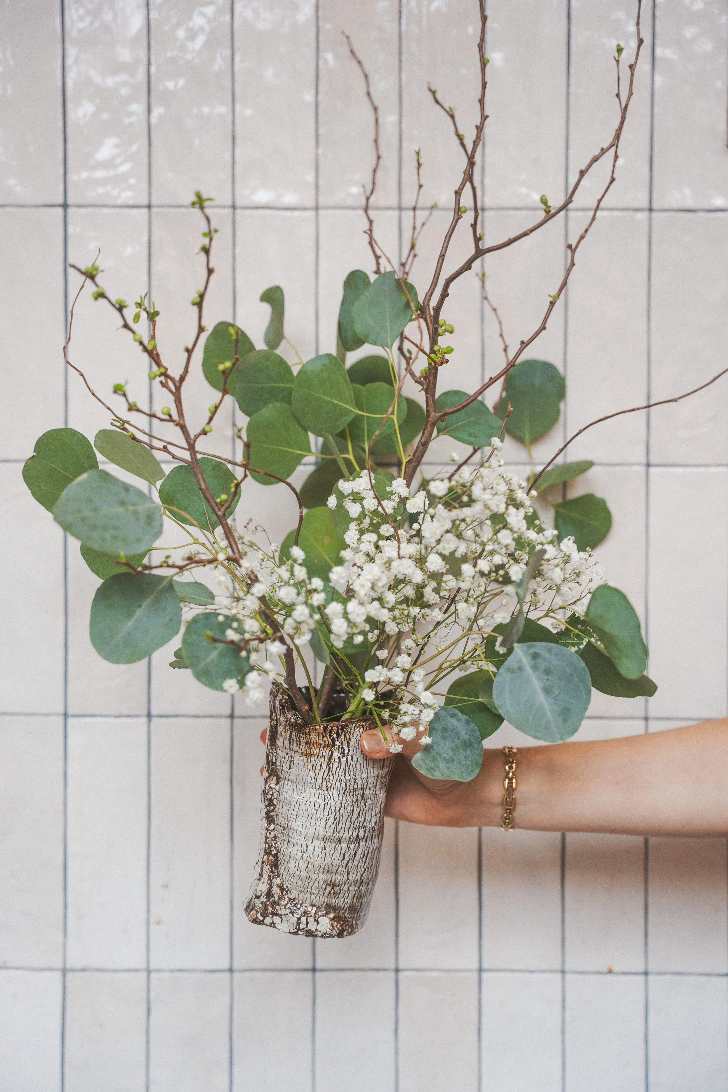 Person holding a bouquet of green leaves, small white flowers, and brown twigs in a rustic bark-covered vase against a tiled background.