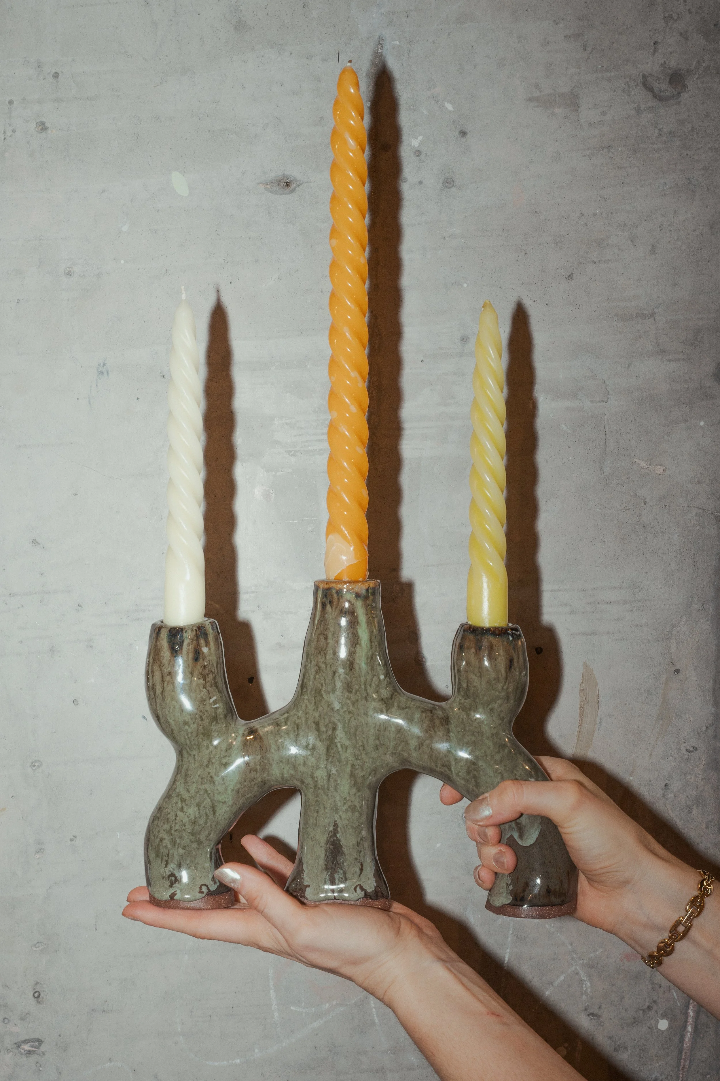 A person is holding a menorah with white, orange, and yellow candles against a concrete wall background.