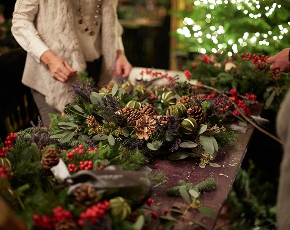 People arranging a festive Christmas wreath made with evergreen branches, pinecones, red berries, and dried flowers.