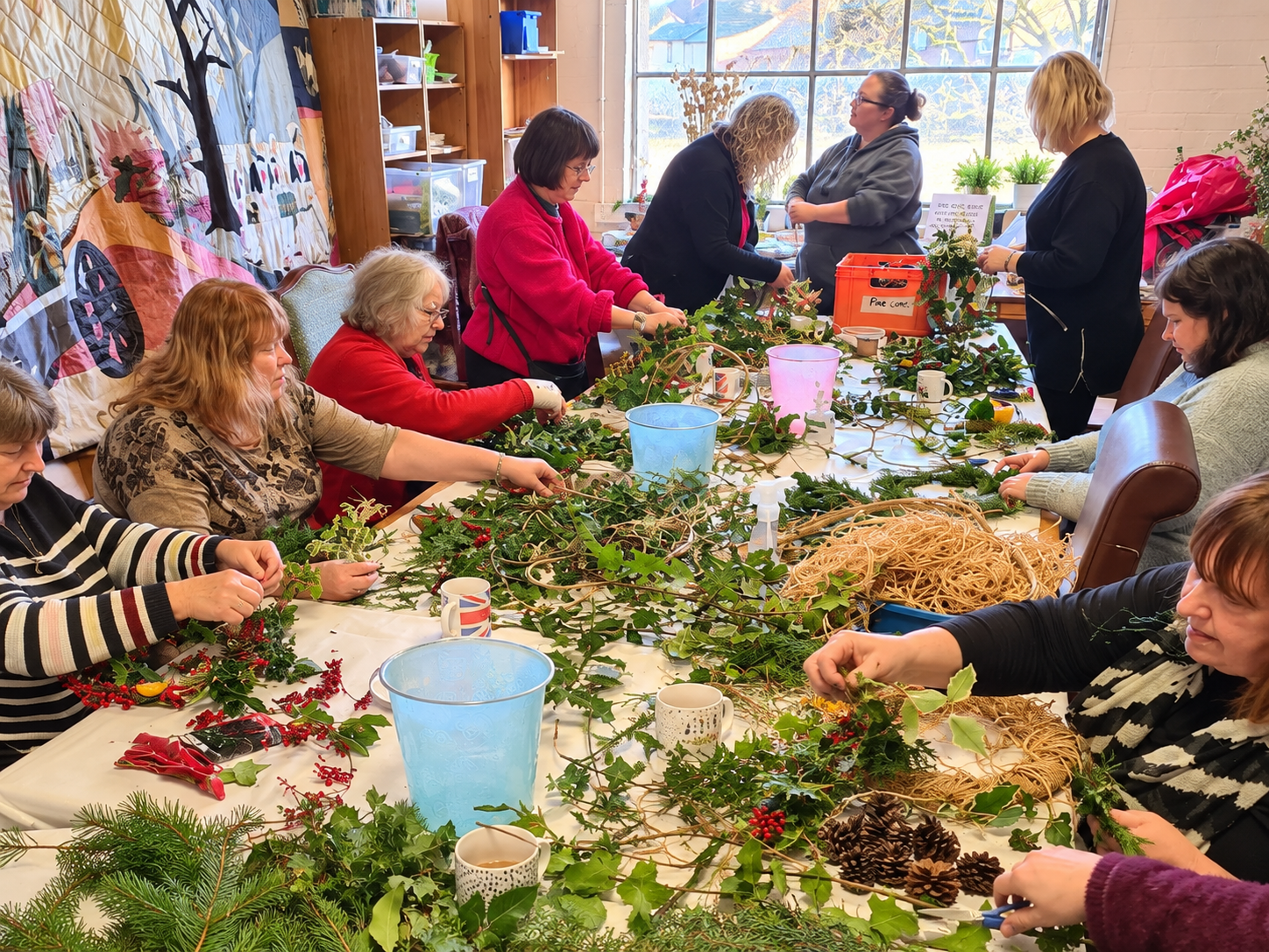 Group of women making festive wreaths from foliage, berries, pinecones, and twine around a workshop table