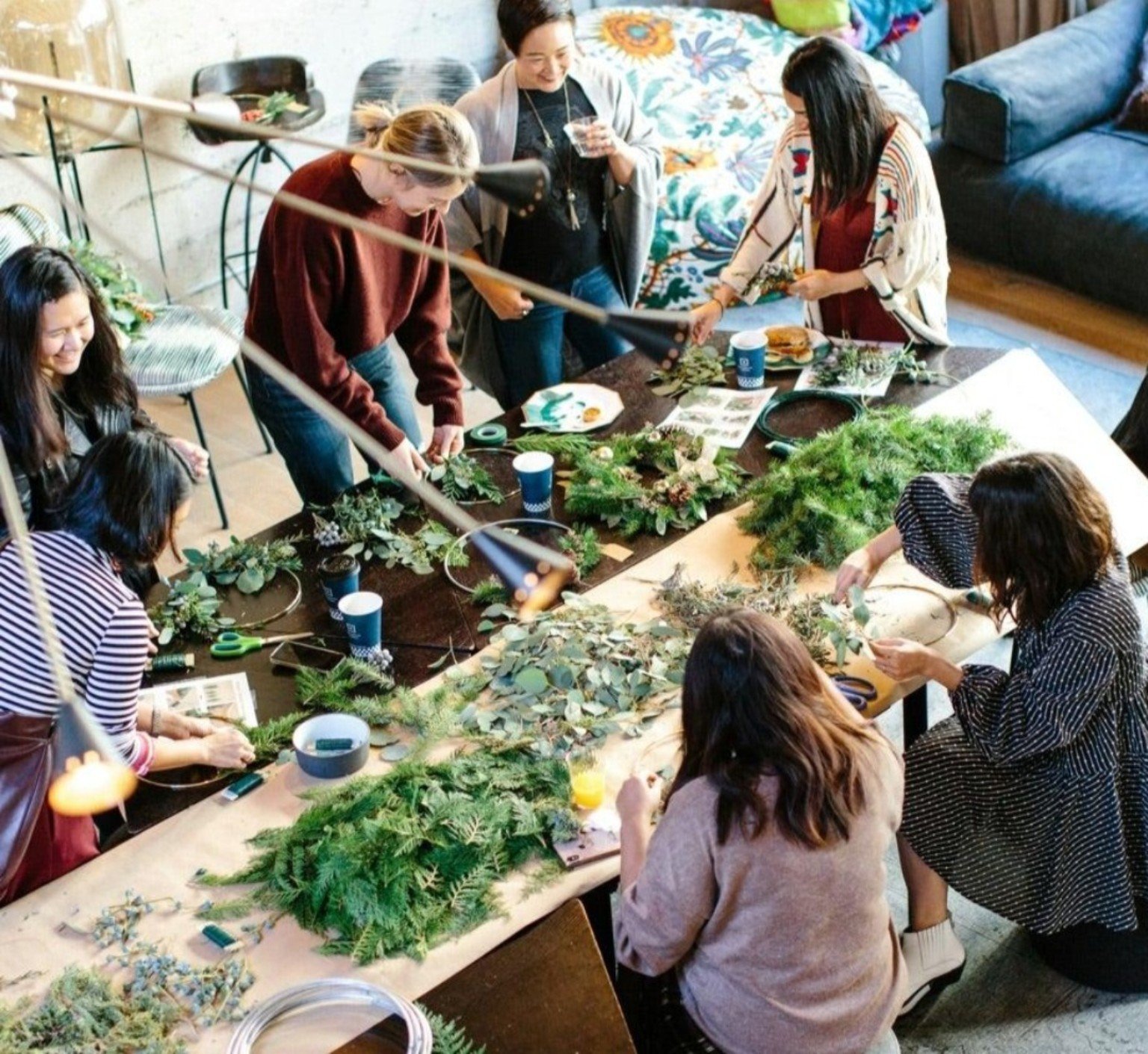 Group of women crafting wreaths and floral arrangements with greenery around a workshop table