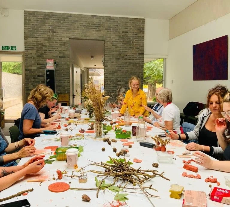 People gathered around a long table making crafts with clay, sticks, leaves, and natural materials in a bright workshop room.