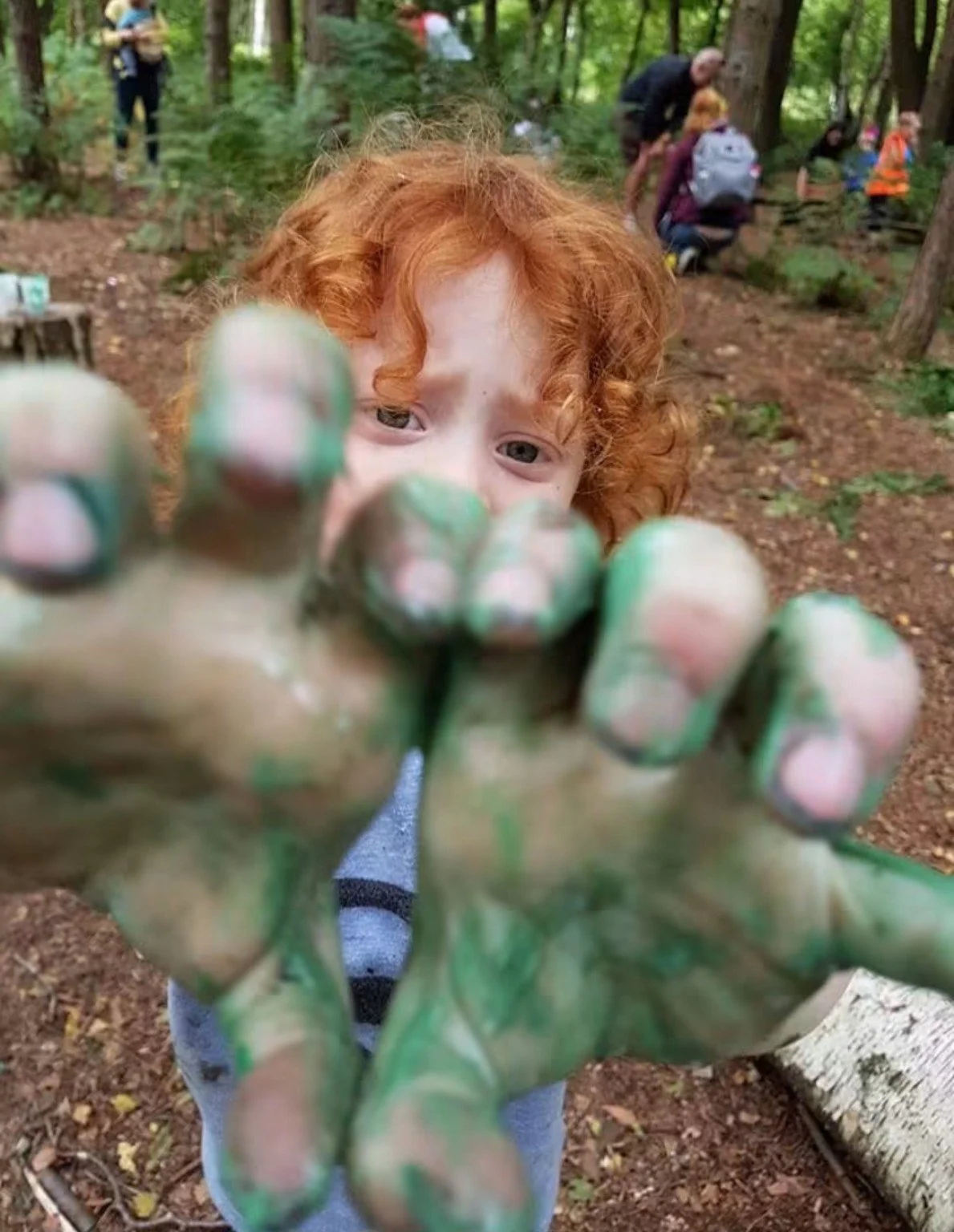 Red-haired child reaching toward the camera with green-painted hands in a wooded outdoor setting, with a group of people in the background