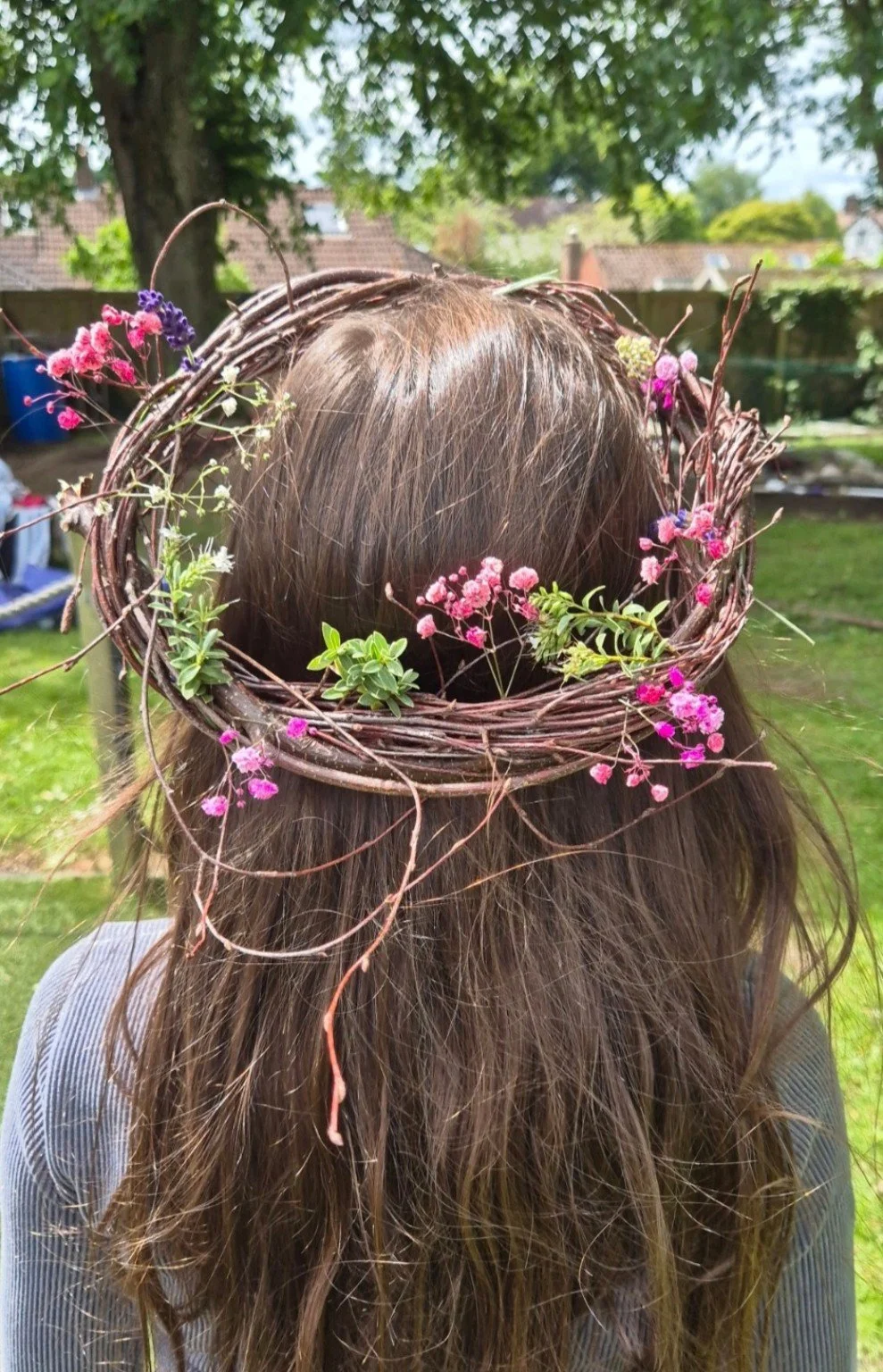 Person wearing a large willow wreath decorated with pink and purple flowers over long brown hair in a garden