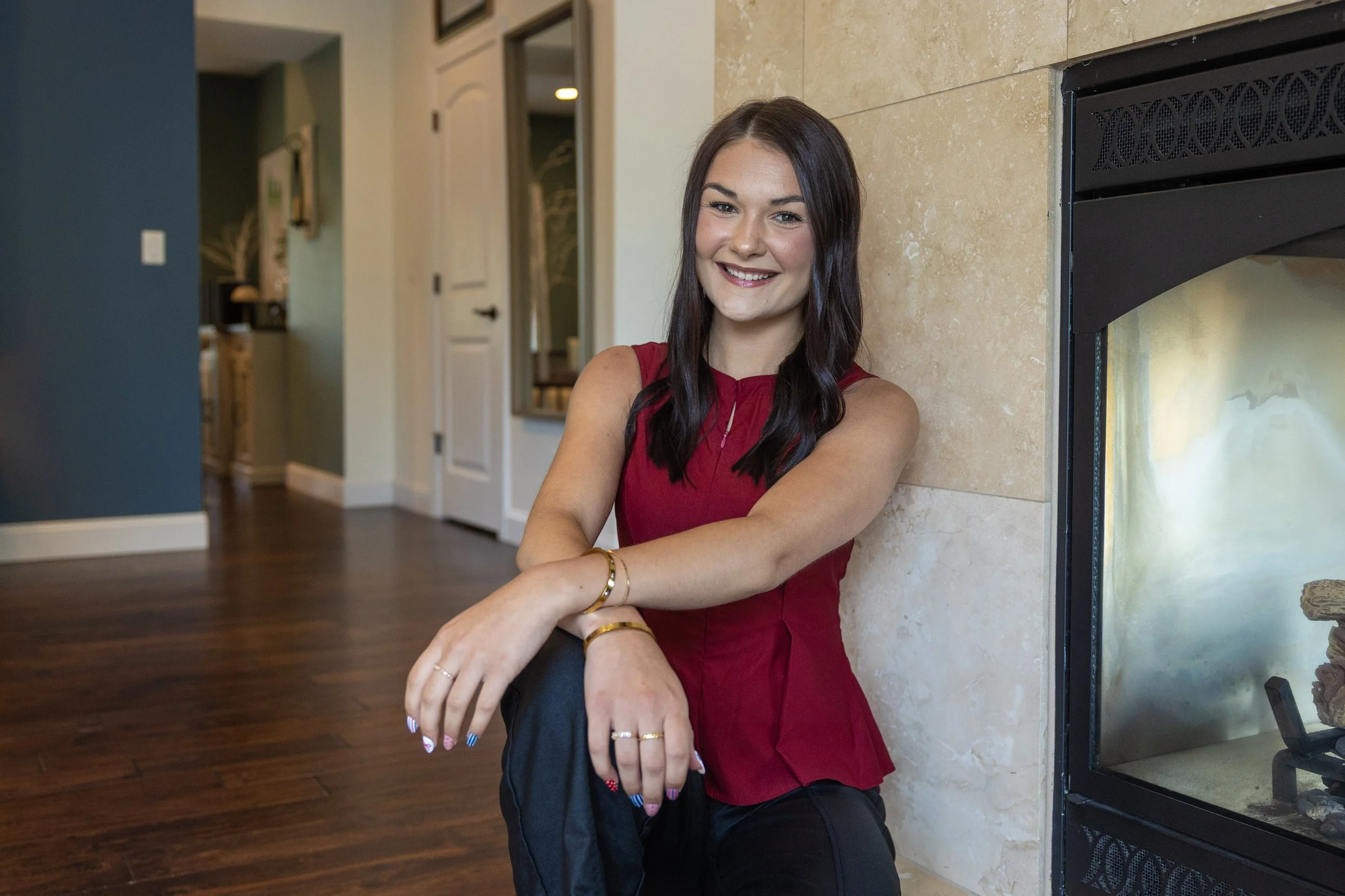 A young woman with long dark hair and a bright smile, wearing a sleeveless red top, stands with her arms resting on her knees, in front of a fireplace in a modern home interior with a wooden floor and a blue accent wall.
