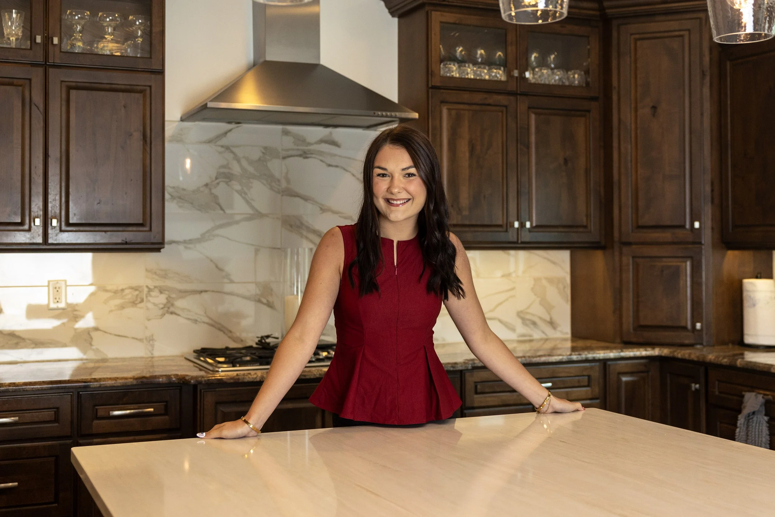 A woman in a red top smiling and leaning on a kitchen counter in a spacious kitchen with dark wooden cabinets and a marble backsplash.