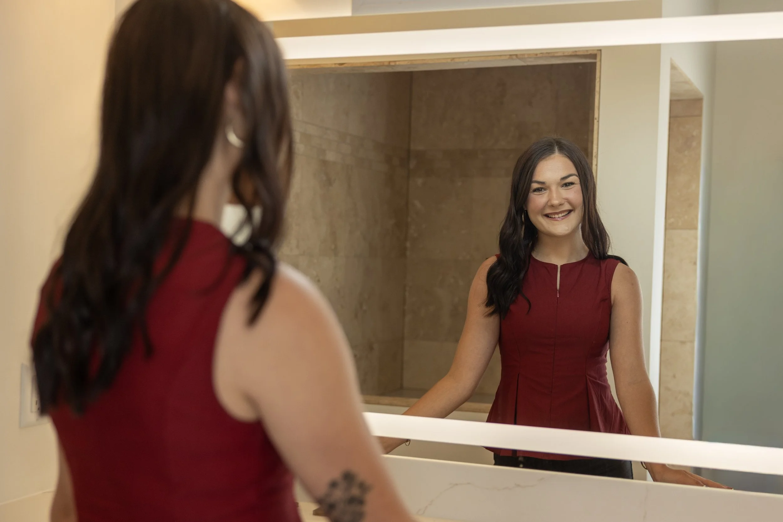A woman with long dark hair wearing a sleeveless red top looking at herself in a bathroom mirror and smiling.