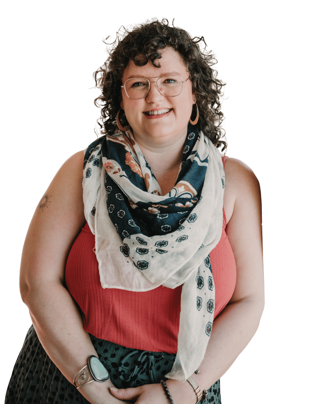 A woman with curly brown hair wearing glasses, a red sleeveless top, a floral scarf, and patterned pants, smiling at the camera.