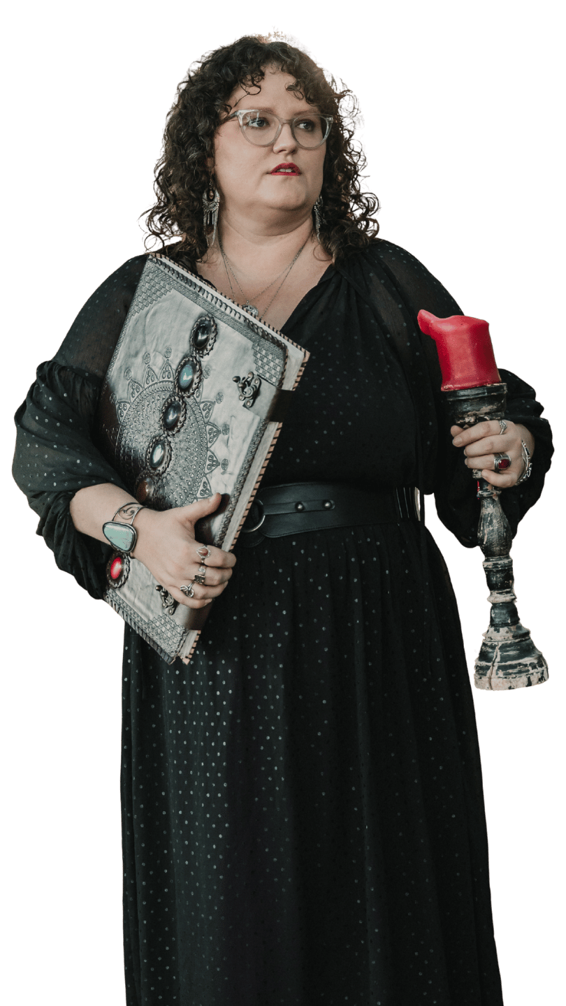 A woman with curly dark hair, glasses, and red lipstick, wearing a black polka dot dress and a sheer black cover-up. She is holding a large, ornate book with gemstones on the cover and a lit red candle in a distressed candleholder.