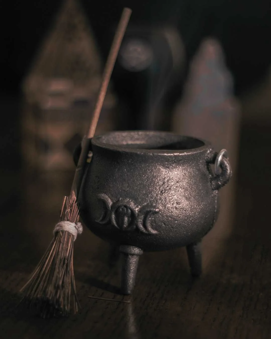 A black cast iron cauldron with three legs, a handle on each side, and a long wooden stirring stick resting inside it. In the background, blurred bottles or containers are visible.